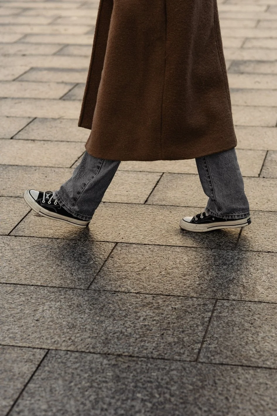 Person wearing a long brown coat, gray jeans, and black Converse sneakers walking on a tiled pavement.