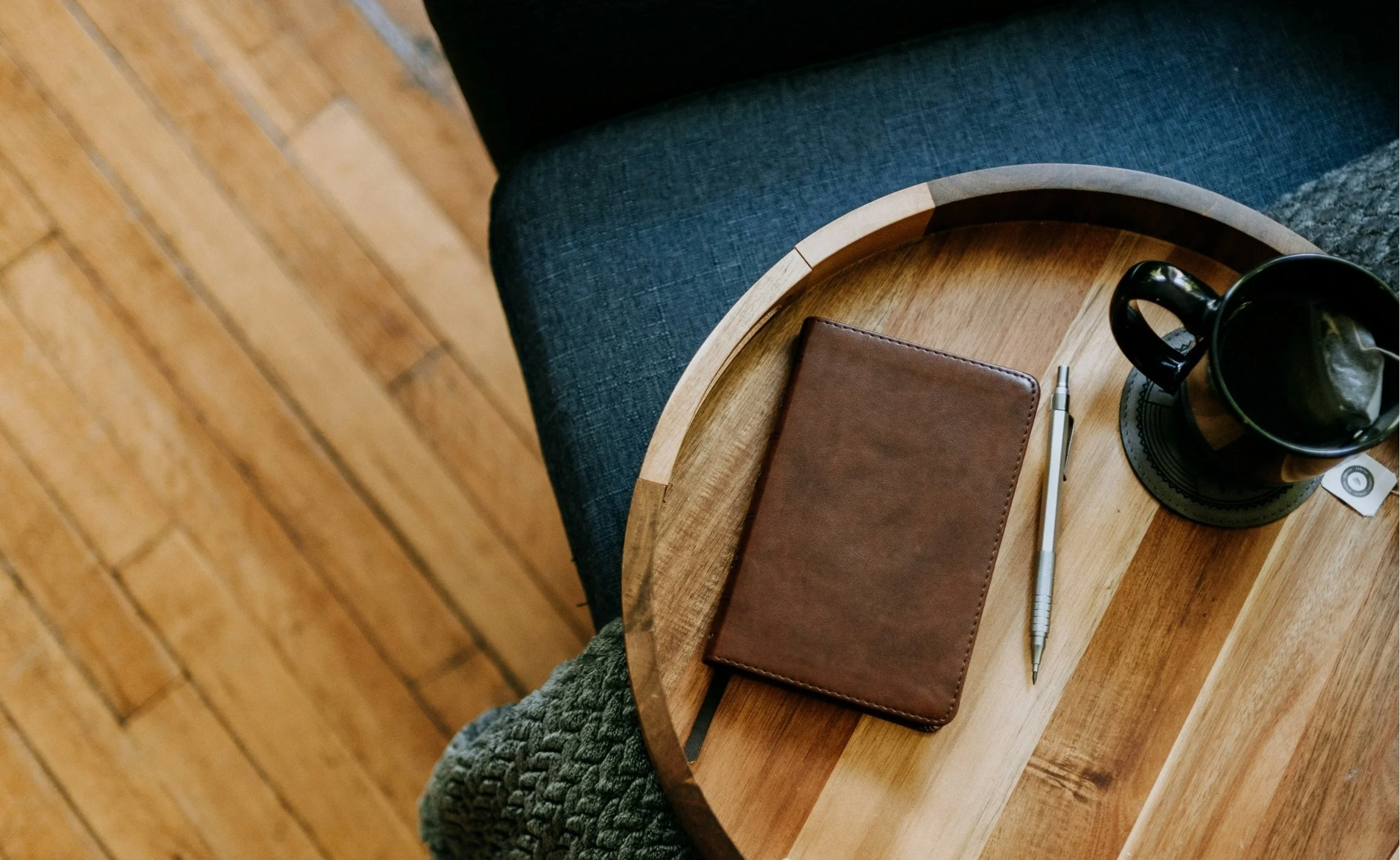 A wooden round side table with a black mug, a brown leather notebook, a white pen, and a small circular object placed on a black doily. A person with a chunky knit sleeve is sitting next to the table on a dark blue upholstered chair with wooden legs in a room with hardwood flooring.