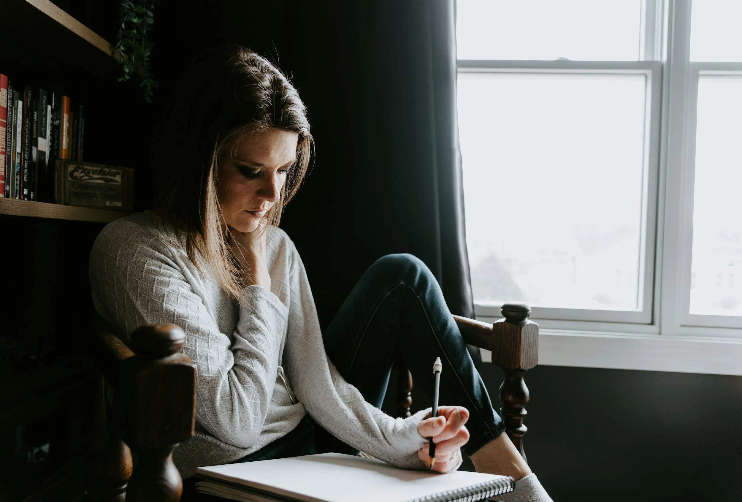 A young woman sitting on a wooden chair near a window, holding a pencil and looking down at an open sketchbook or notepad, in a room with a bookshelf and green plant in the background.