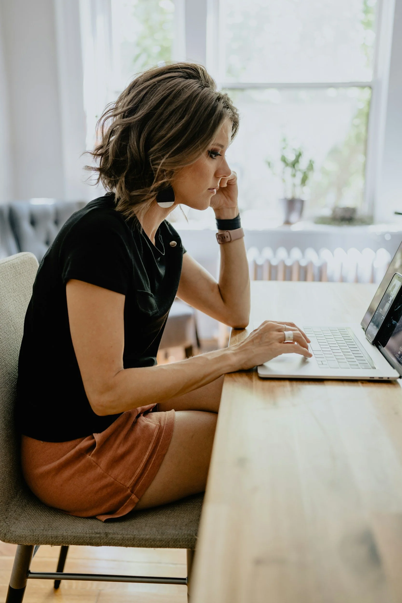 Woman with short wavy hair wearing earrings, a black shirt, and shorts, sitting at a wooden table working on a laptop in a bright room with large windows and potted plants.