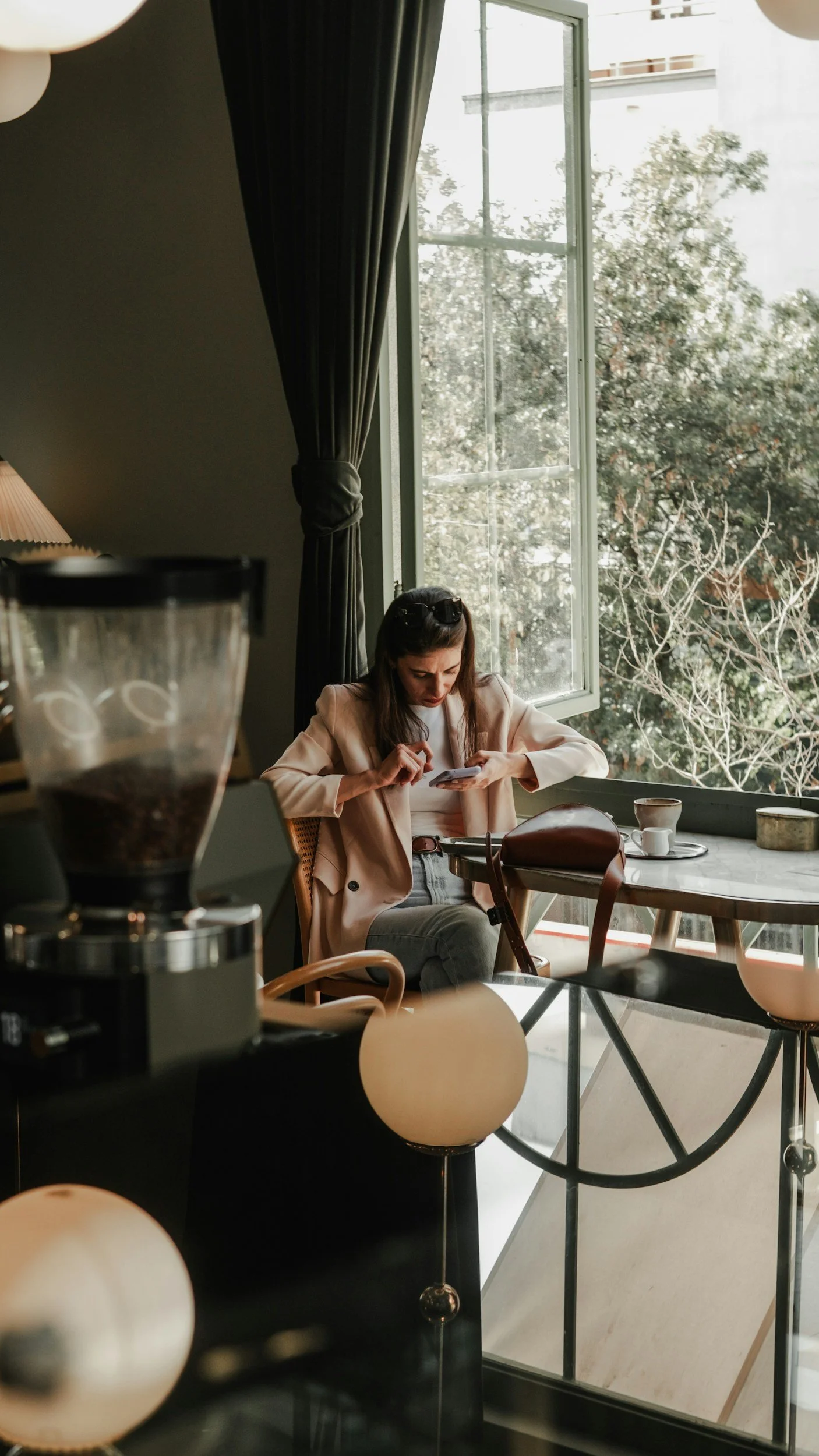 A woman sitting at a café table, looking at her phone near a window with trees outside, coffee cups on the table, and coffee grinder in the foreground.