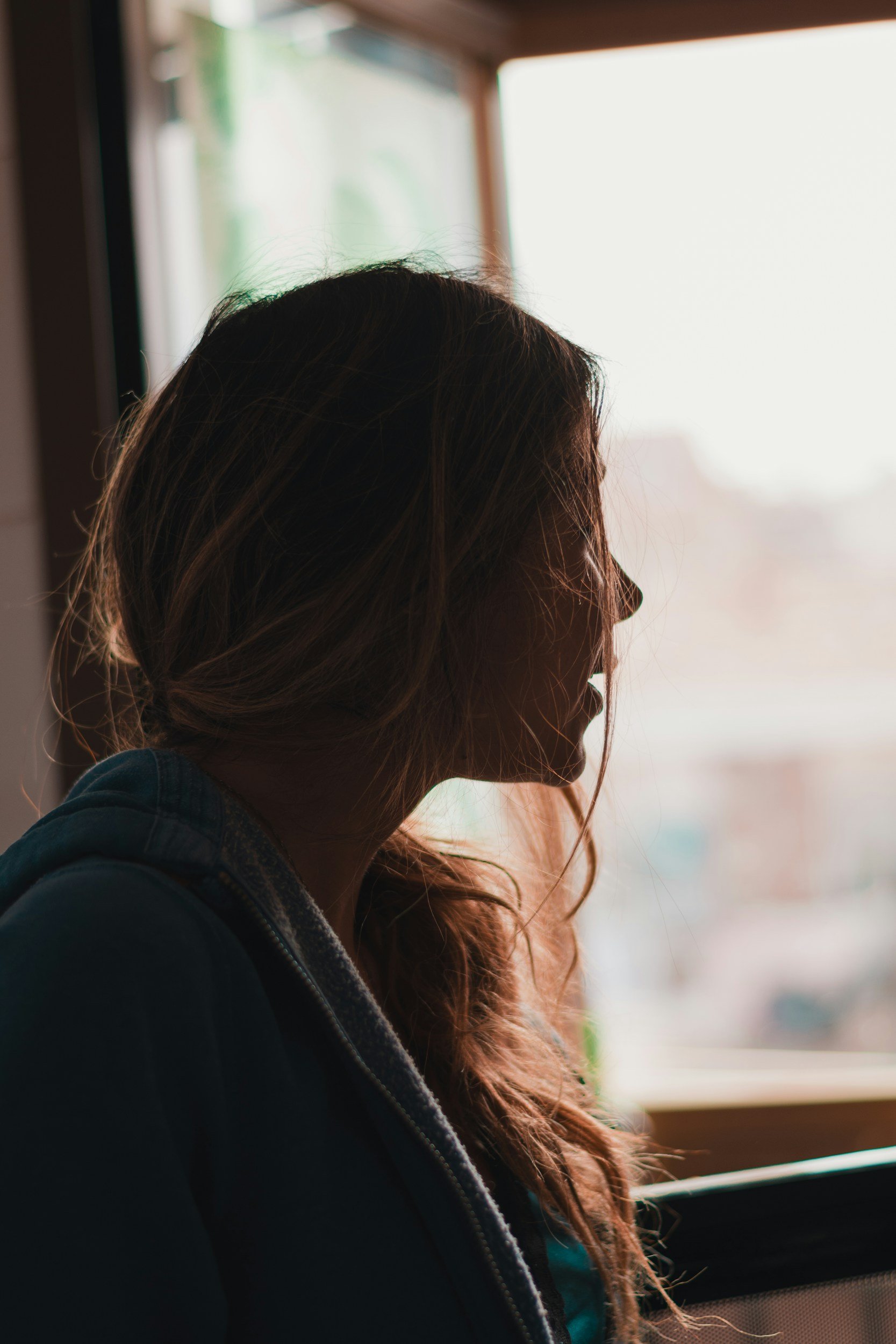 A woman with long brown hair sitting by a window, looking outside on a bright day.