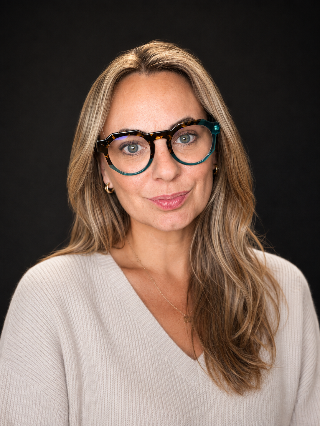 A woman with light brown, wavy hair wearing large tortoiseshell glasses, gold earrings, a gold necklace, and a light beige sweater against a black background.