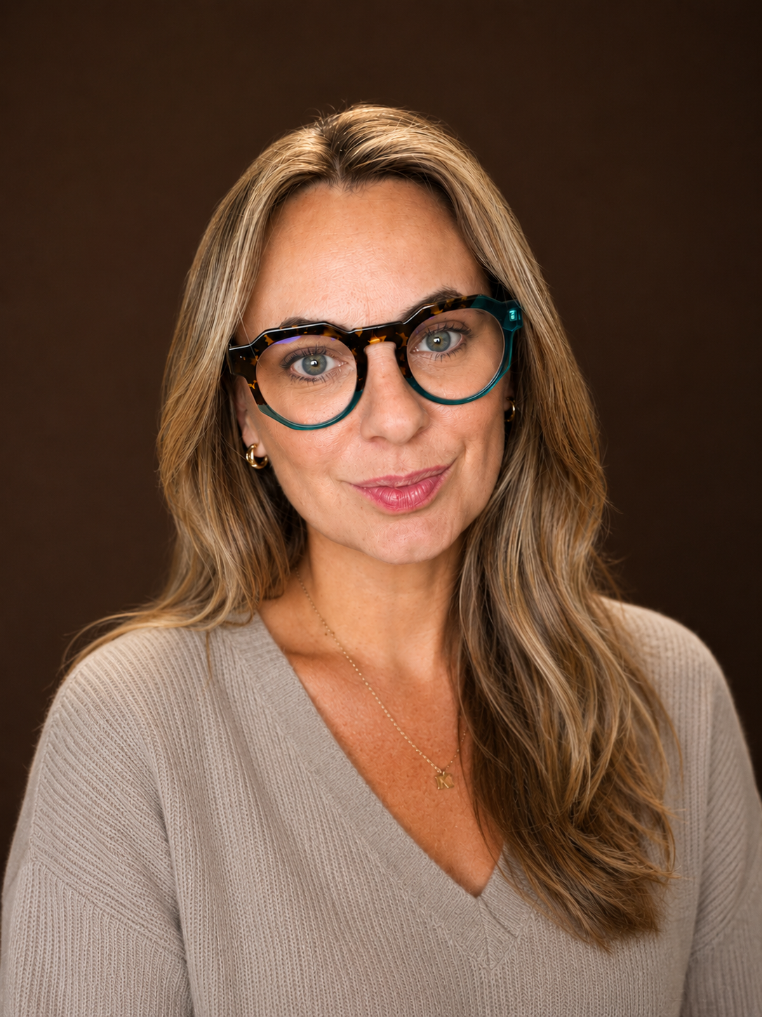 A woman with wavy blonde hair wearing large tortoise shell and turquoise glasses, gold hoop earrings, a delicate gold necklace with a letter 'K', and a beige V-neck sweater, smiling softly against a dark brown background.