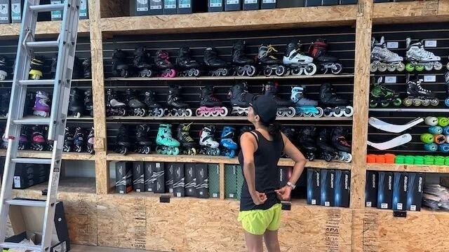 A young woman wearing a black tank top, bright yellow shorts, and a black cap looks at a display of rollerblades in a skate shop. The rollerblades are organized on wooden shelves and come in various colors. There are also skateboards stored on the right side of the shelf. A silver ladder leans against the shelving unit on the left side.
