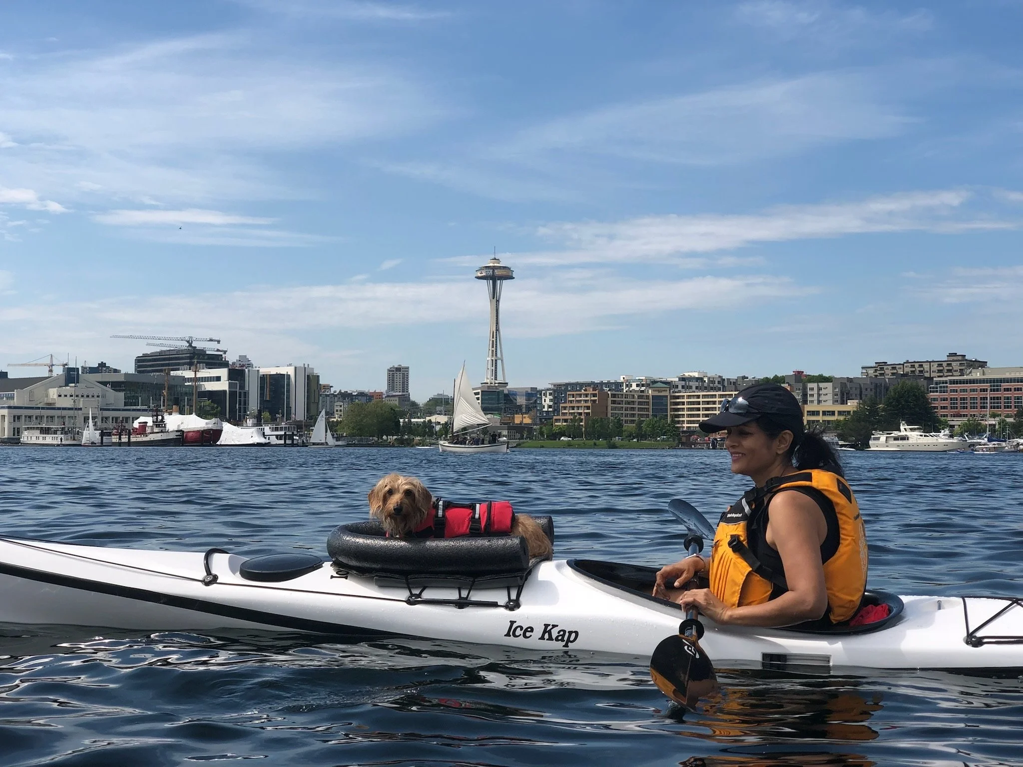 A woman in a yellow life jacket and black cap smiles while kayaking on a body of water, with a dog sitting on the kayak's front. In the background, the Seattle skyline with the Space Needle and boats on the water are visible under a partly cloudy sky.