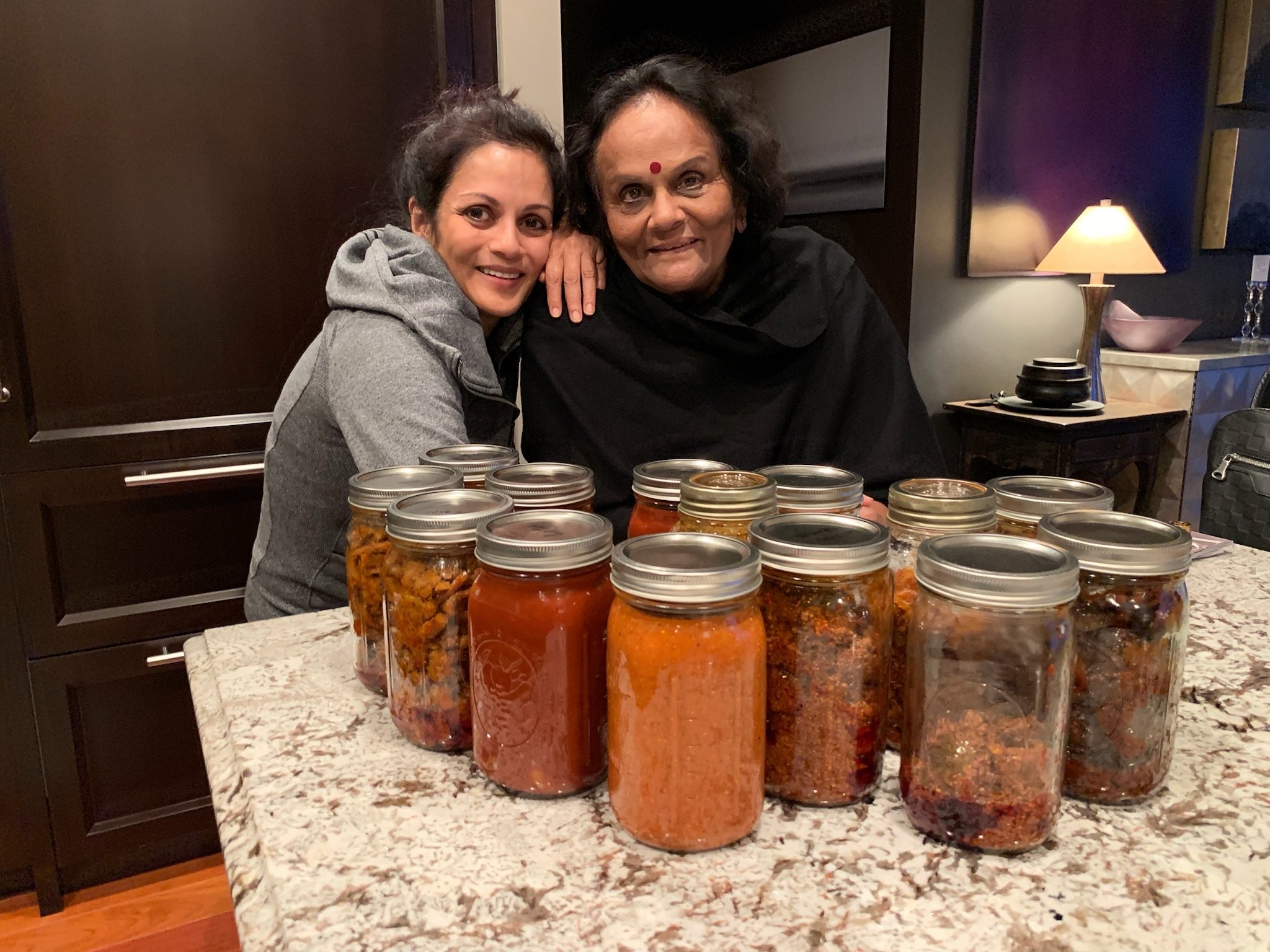 Two women smiling, one older and one younger, sitting at a table with numerous jars of preserved food. The setting appears to be a cozy kitchen or dining area with a granite countertop and a lamp in the background.