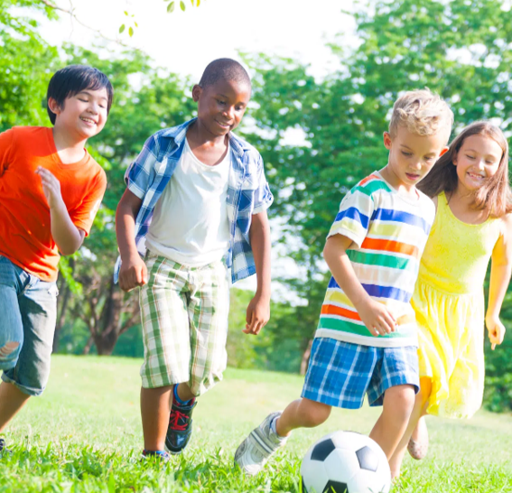 Children playing soccer outdoors on a grassy field with trees.