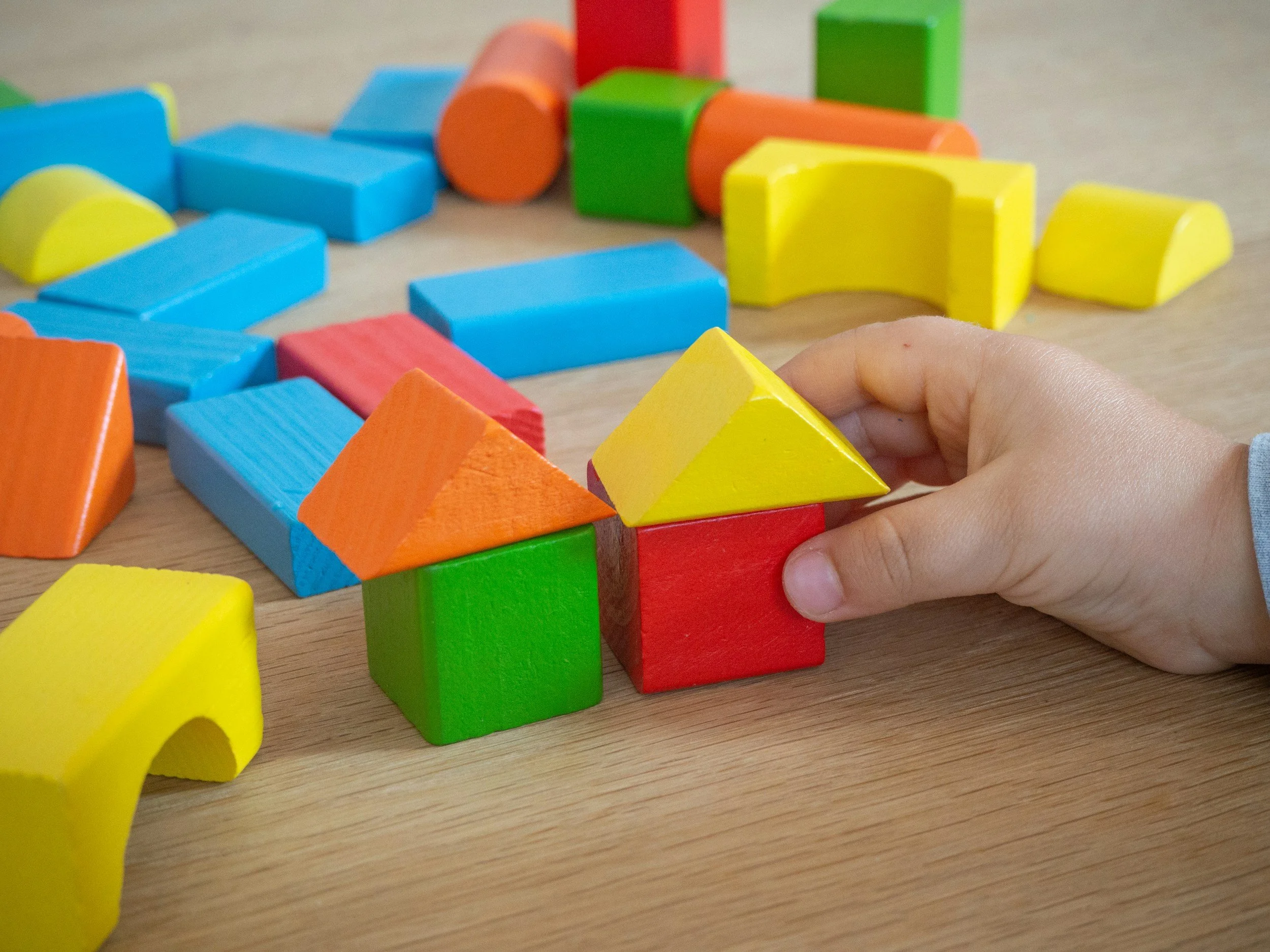 Colorful wooden building blocks arranged on a wooden surface, with a child's hand playing with a yellow block.