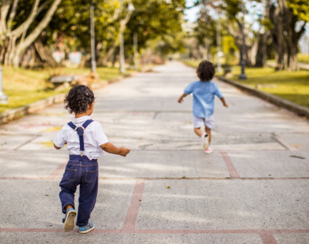 Two young children, one in overalls and the other in a blue shirt, running on a paved road in a park with trees and greenery.