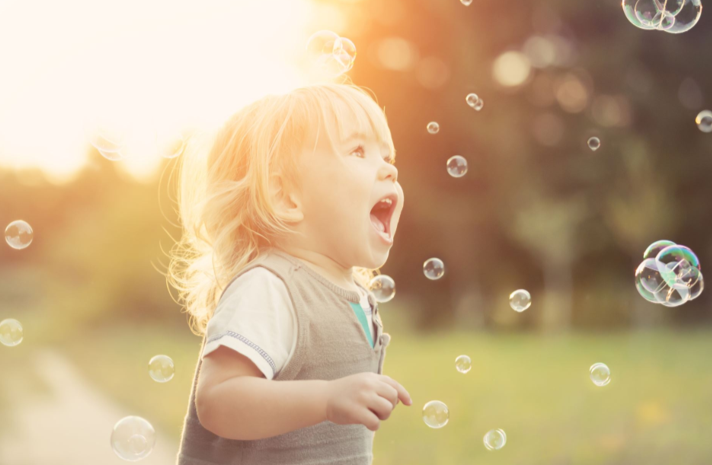 A young girl with blonde hair playing with soap bubbles outdoors during sunset.