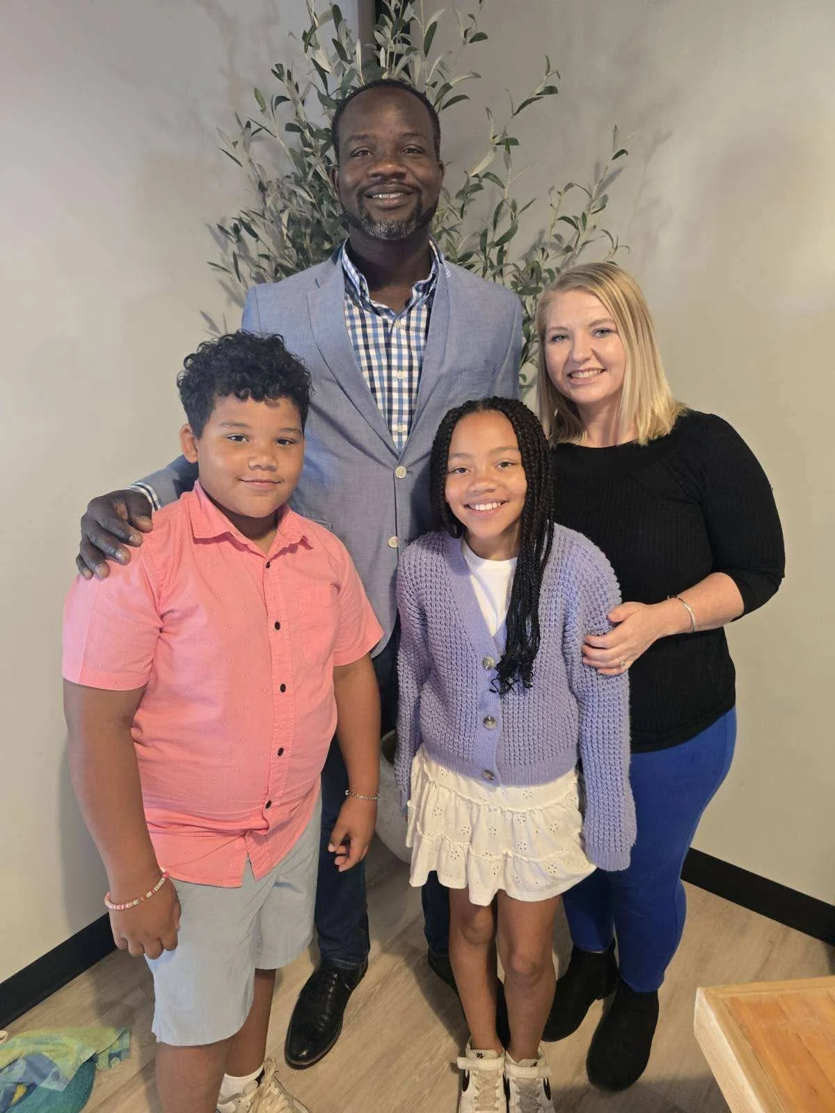A diverse family group consisting of a man, a woman, and three children smiling and posing together indoors, with a large plant in the background.