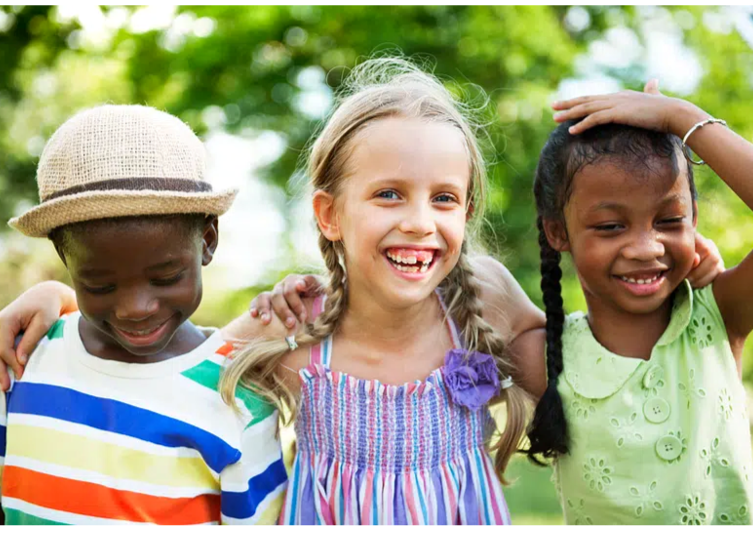 Three young girls smiling outdoors with green trees in the background, one wearing a straw hat and colorful striped shirt, another in a patterned dress, and the third in a green top with braids.