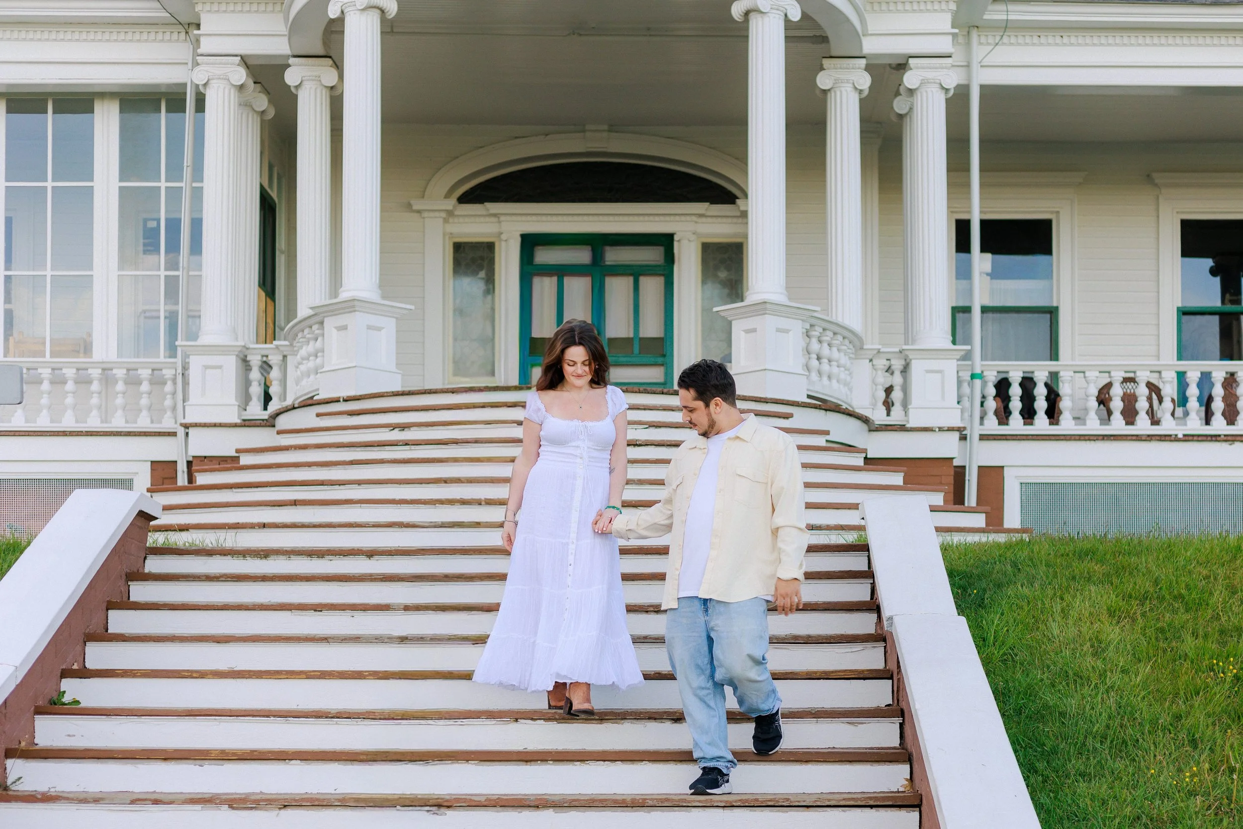 A couple holding hands and walking down the steps of a large, classic white house with columns and a porch. The woman is wearing a white dress, and the man is wearing a light jacket and jeans.