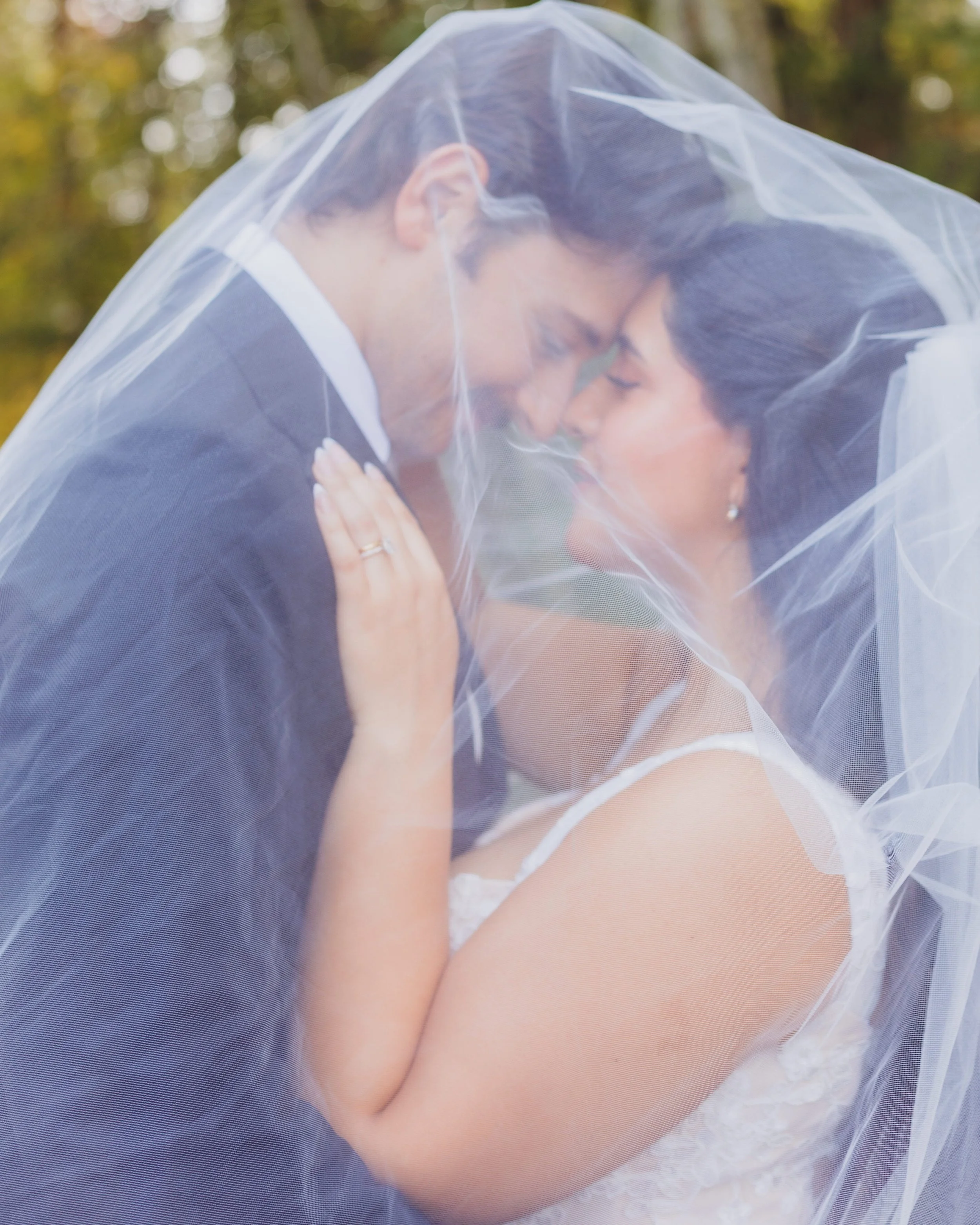 A bride and groom embrace under a wedding veil in an outdoor setting with trees in the background, creating a romantic moment.