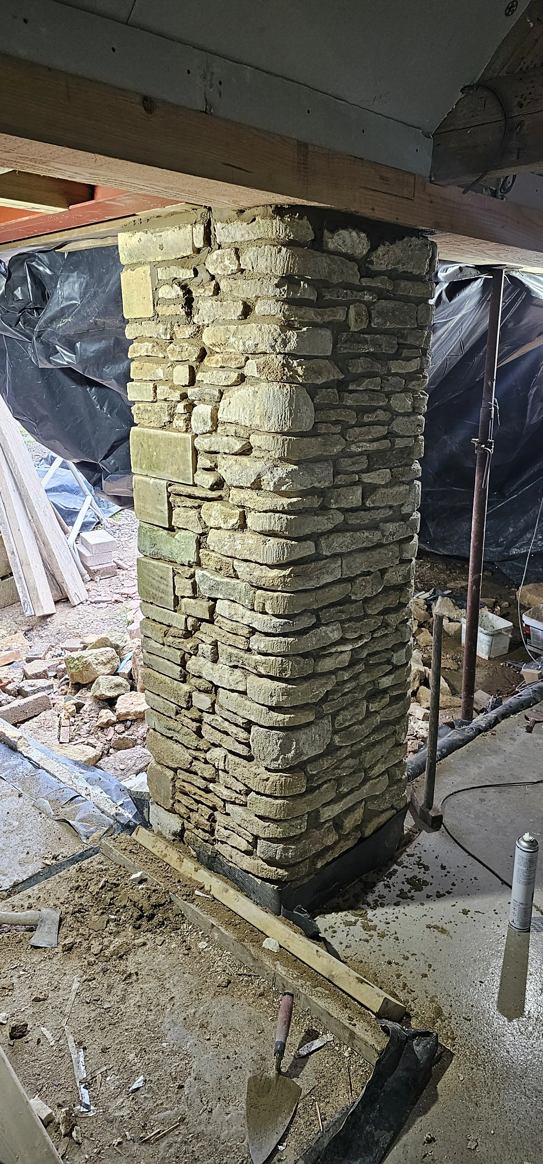 Indoor construction site featuring a stone chimney with irregular stones, wooden beams overhead, construction tools, debris, and black plastic sheeting in the background.