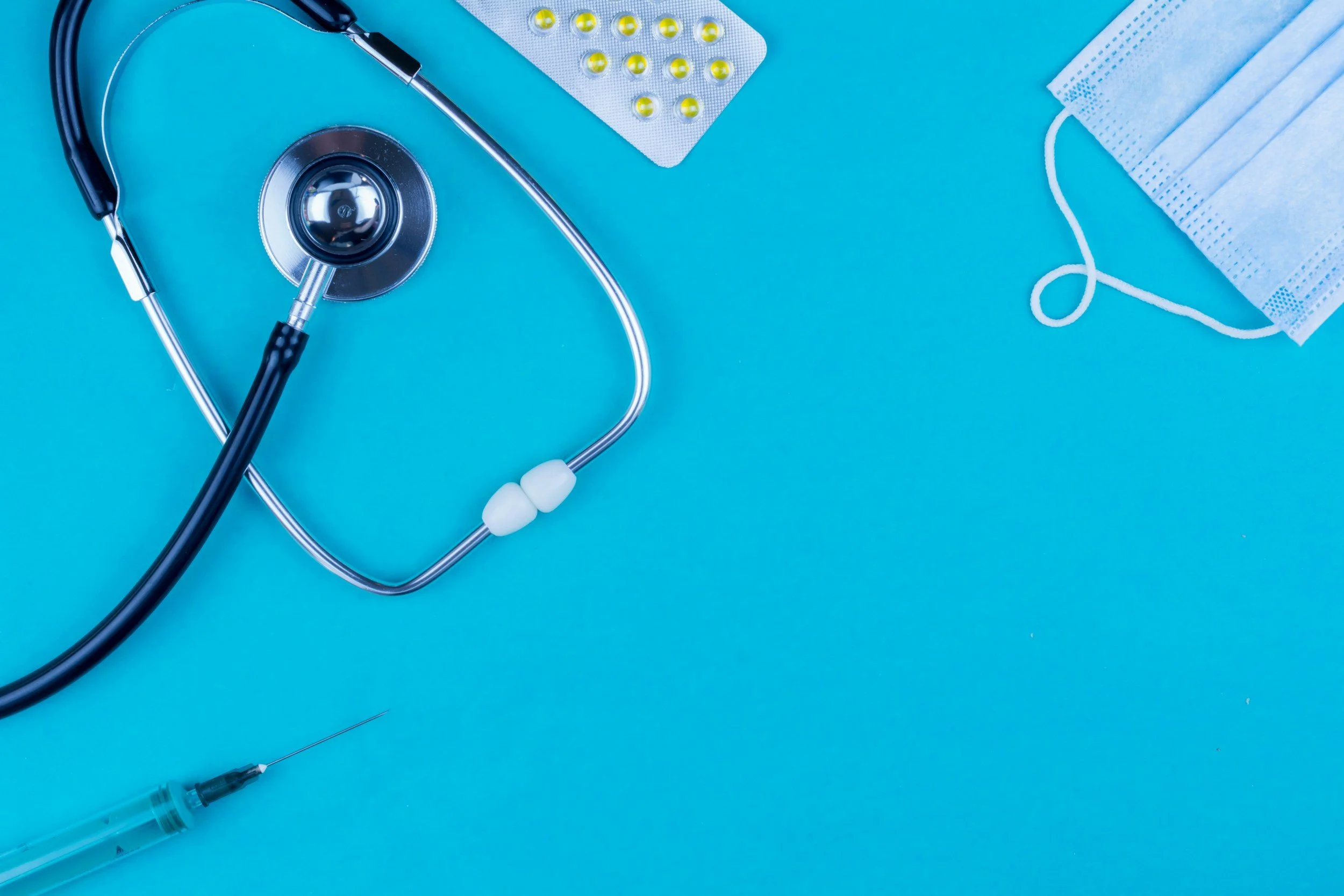 Medical equipment including a stethoscope, blister pack of yellow pills, face mask, and syringe on a blue background.