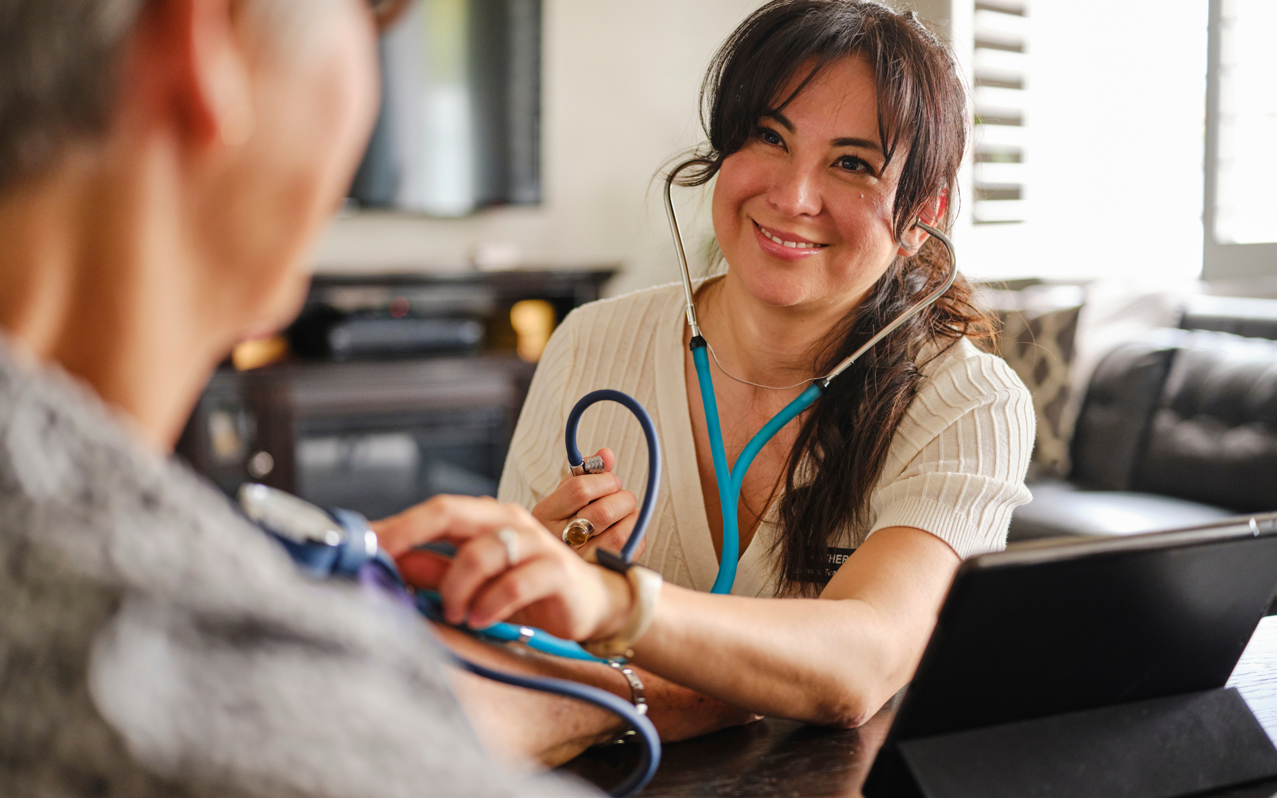 A healthcare professional, smiling, wearing a beige shirt and using a stethoscope to check a patient's blood pressure, with a bright, modern room and large windows in the background.