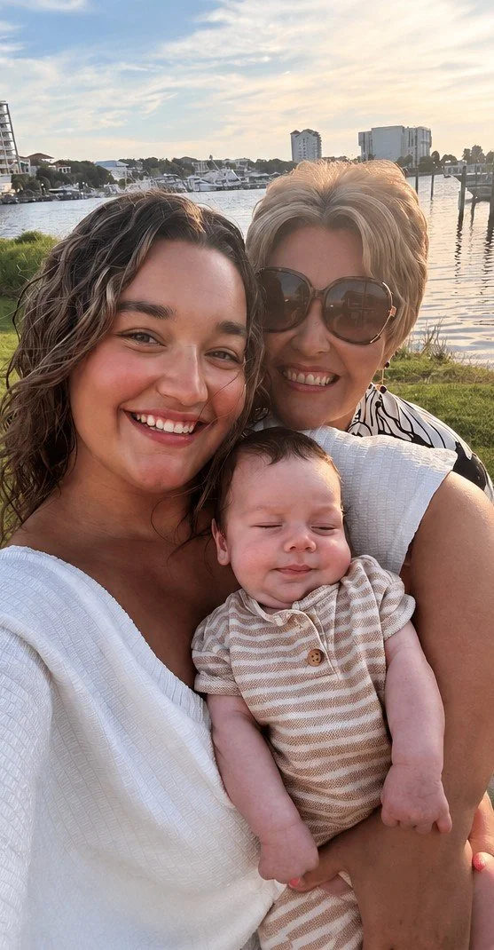 Three people, two women and a baby girl, smiling outdoors near a body of water with boats, buildings, and a partly cloudy sky in the background.