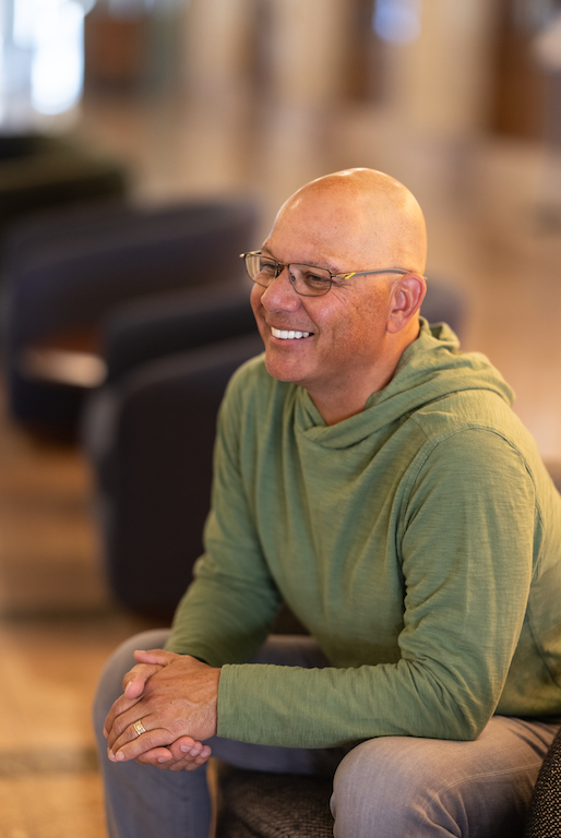 A smiling man with glasses and a bald head, wearing a green hoodie, sitting on a chair in a well-lit indoor space.