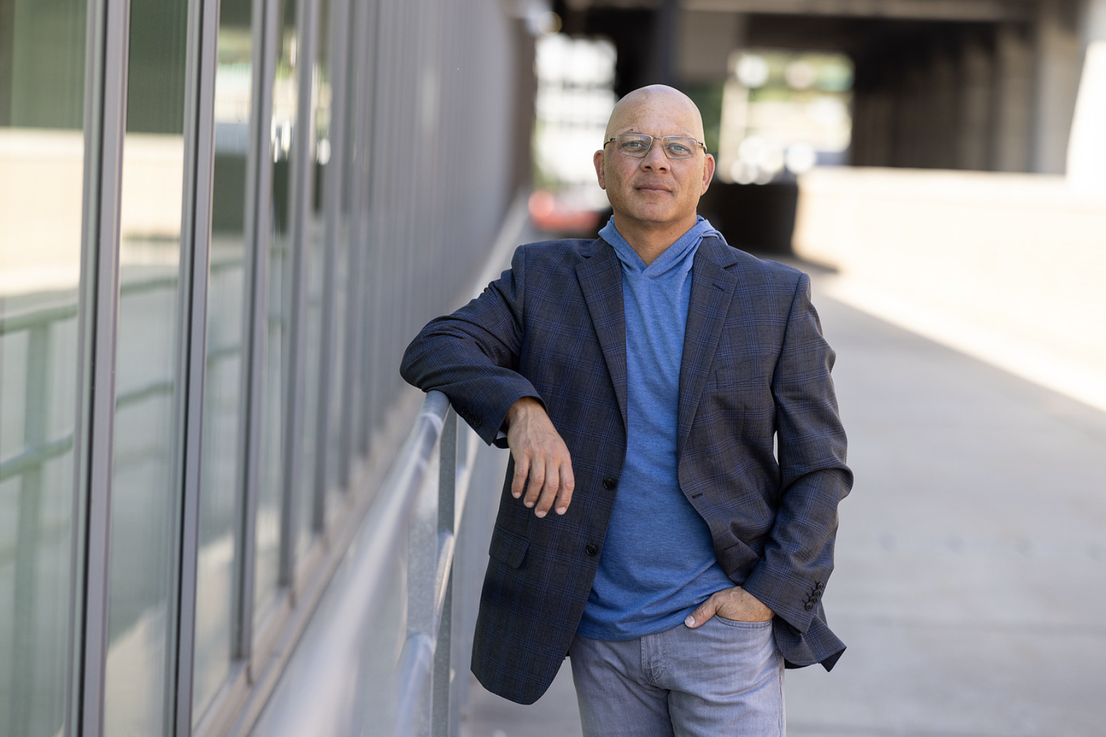 A bald man with glasses wearing a blue hoodie, a checked blazer, and gray pants leaning against a railing outdoors.
