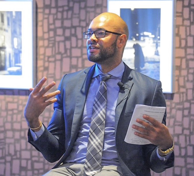 Man with glasses in a business suit giving a presentation or speaking, holding papers, in a room with framed photos on the wall.
