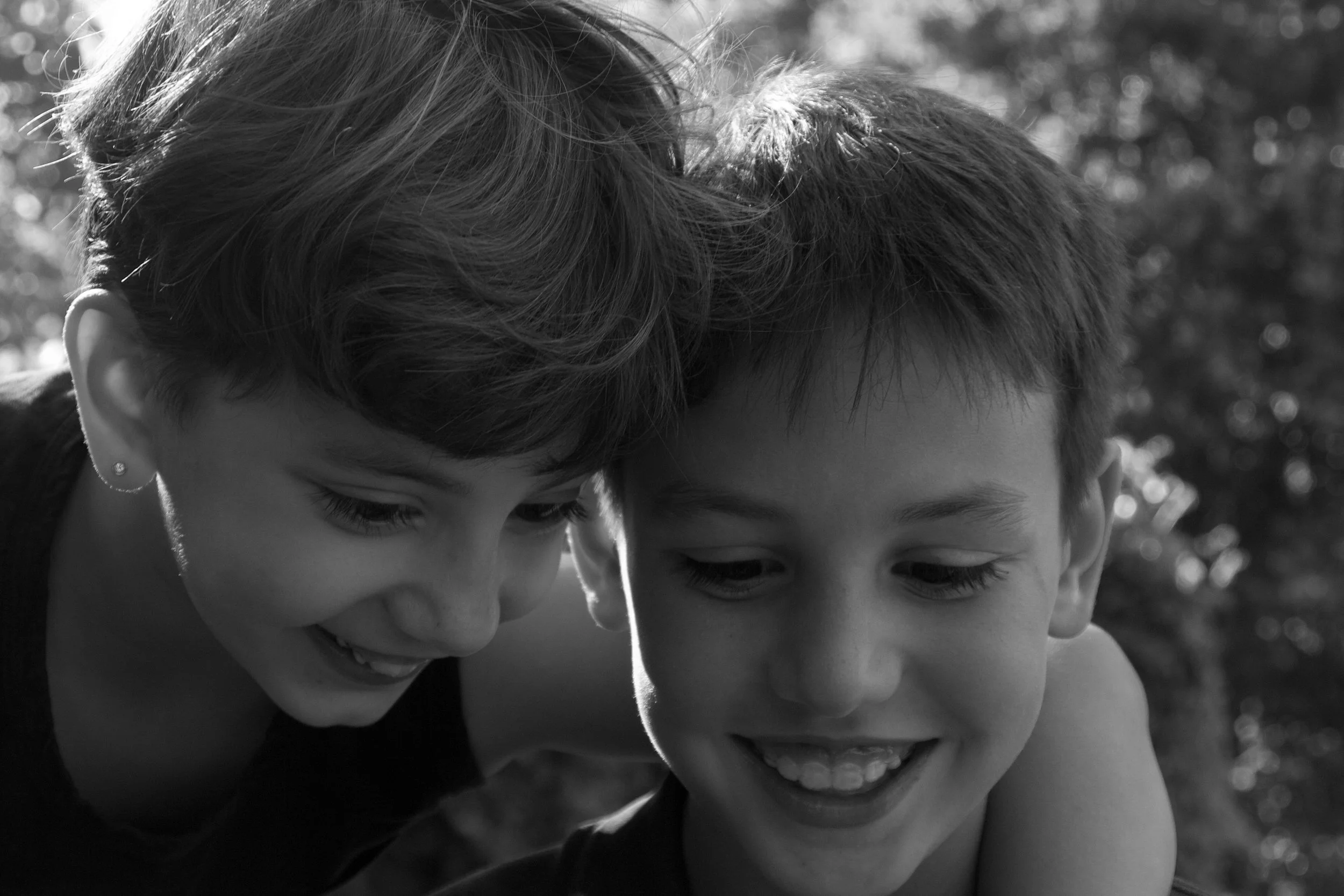 Natural family portrait of two siblings sitting together outdoors, photographed in soft natural light with relaxed, spontaneous expressions.