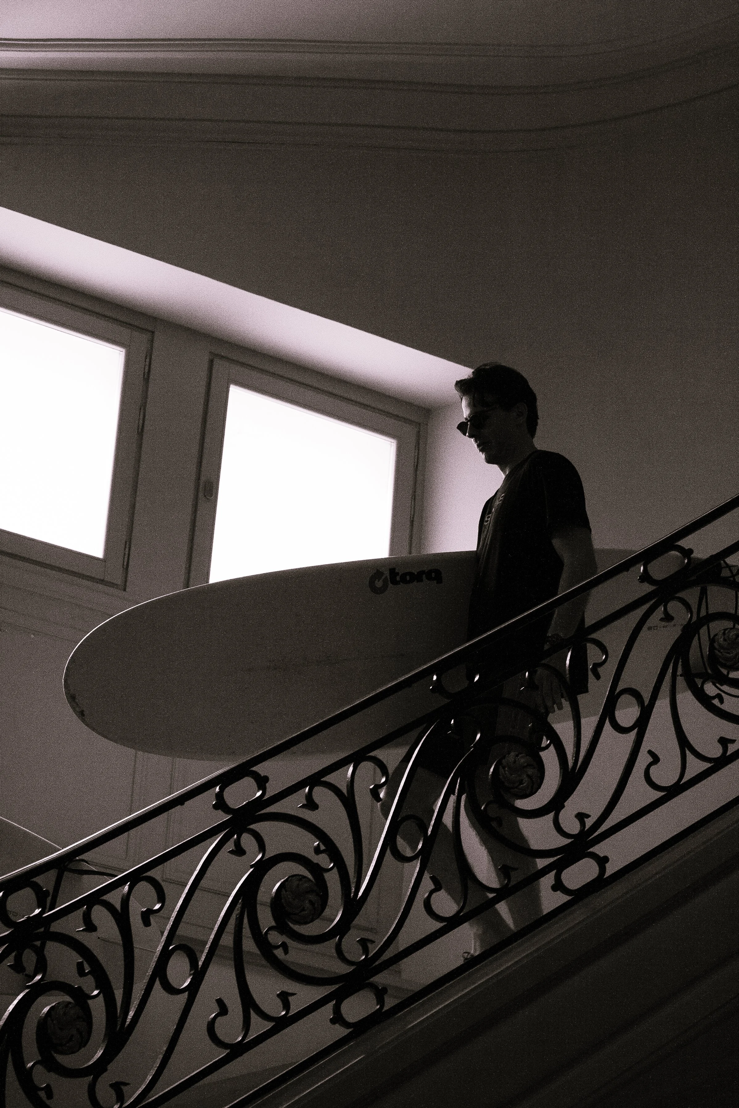 Black-and-white editorial lifestyle photograph of a surfer carrying his board through the grand interior of a luxury hotel in Biarritz.