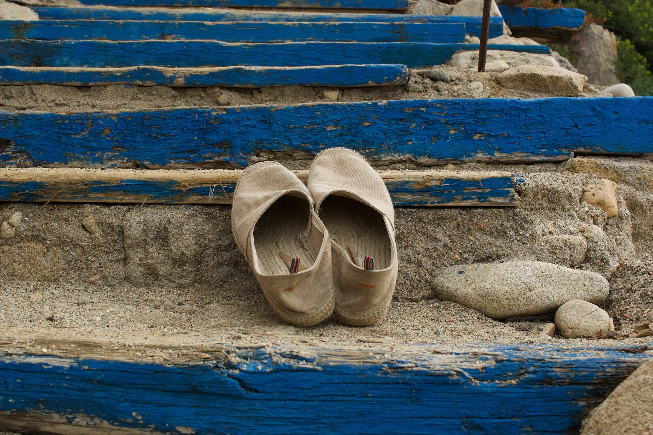 Neutral canvas espadrilles on weathered blue steps by the Mediterranean, capturing the relaxed summer styling of the Pietro SS26 fashion story.