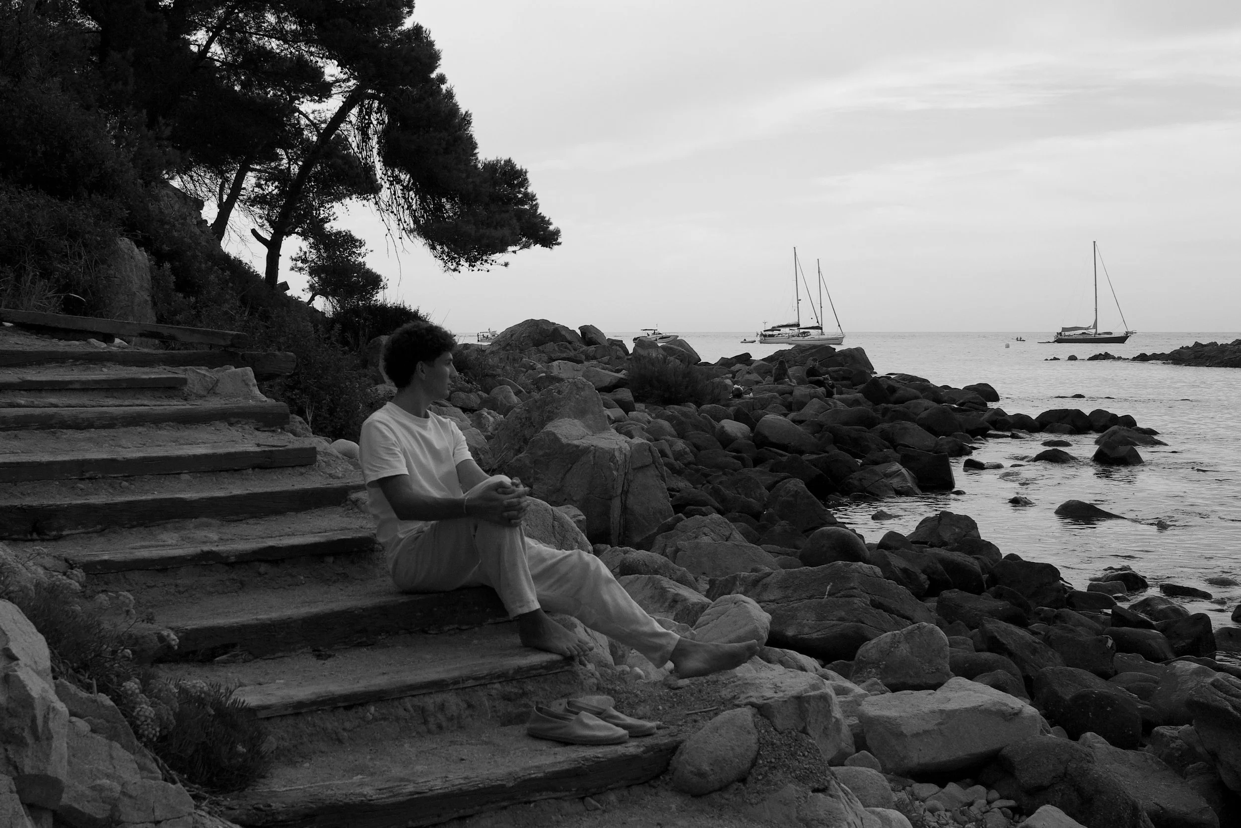 Black-and-white men’s fashion portrait on the Costa Brava, featuring a white cotton T-shirt and relaxed linen trousers in an effortless Mediterranean setting.