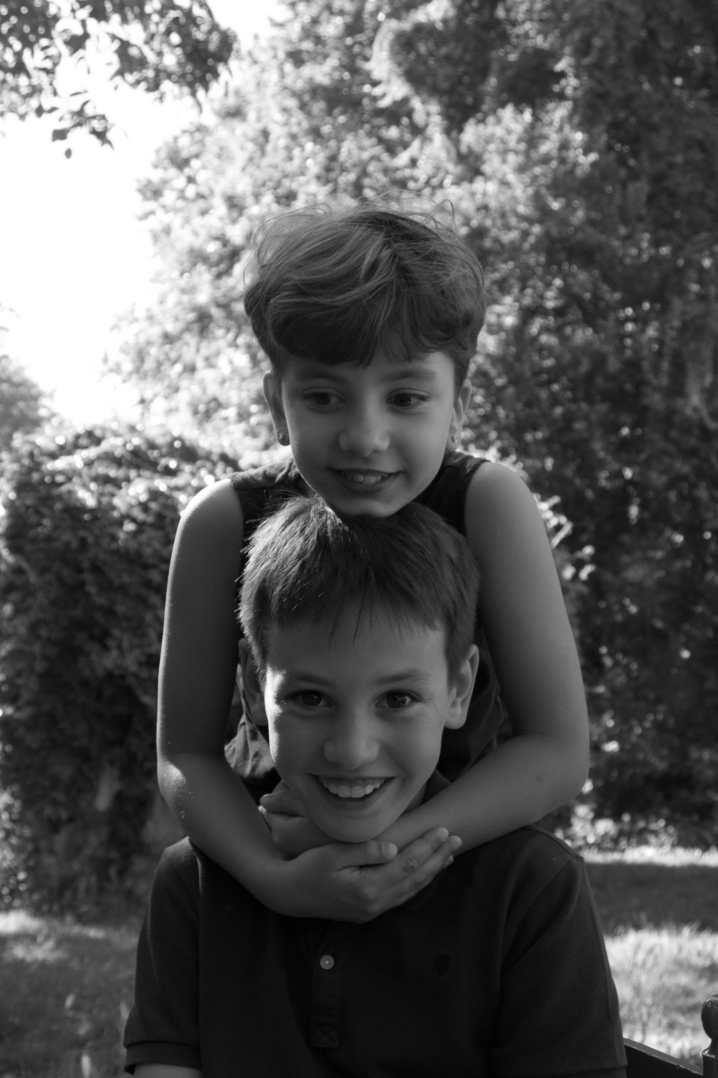 Candid black-and-white sibling portrait outdoors, capturing two children playing together with relaxed expressions and natural smiles.