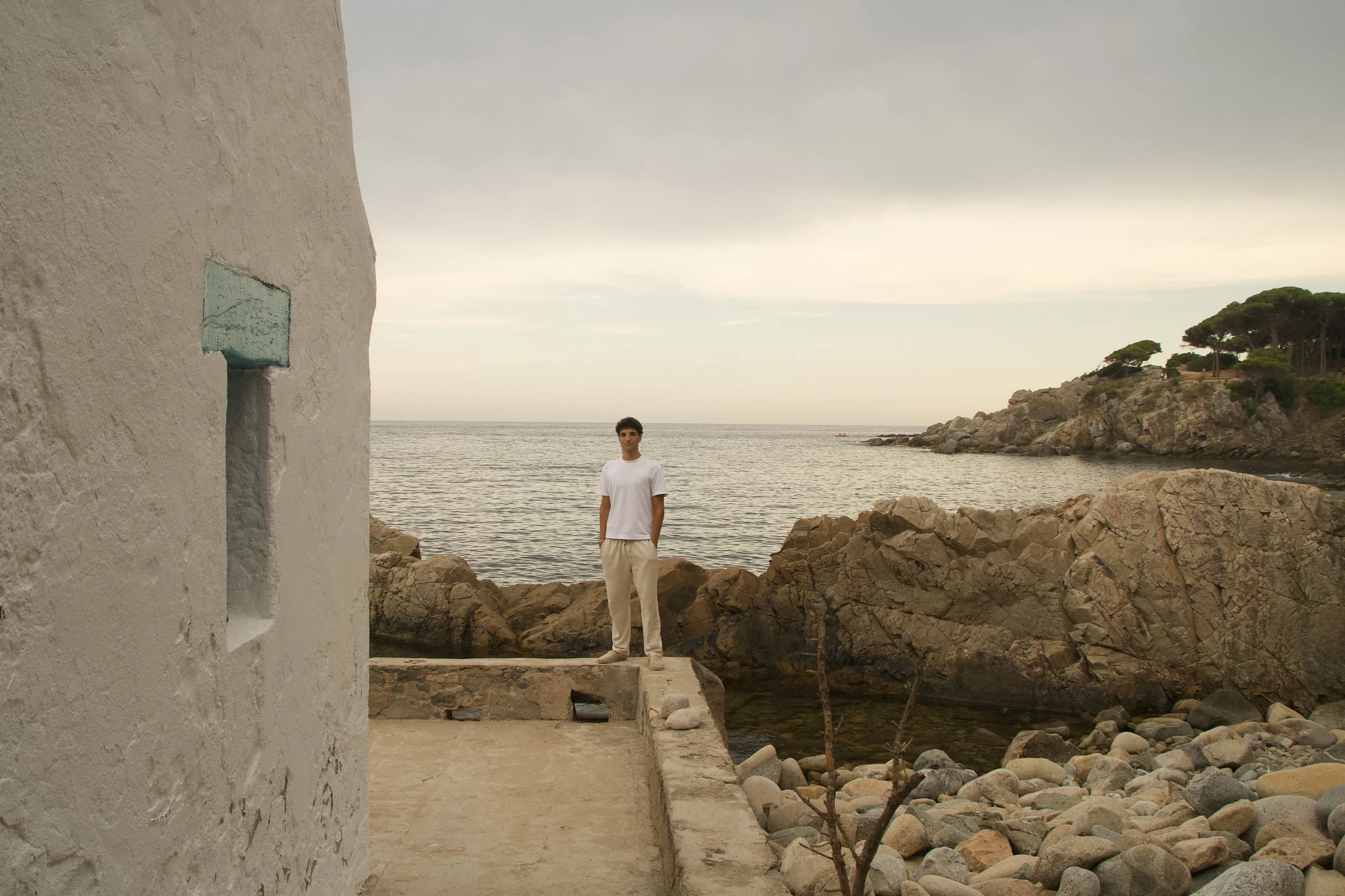 Relaxed Mediterranean fashion portrait of a young man in a white cotton T-shirt and linen trousers walking beside the sea on the Costa Brava.