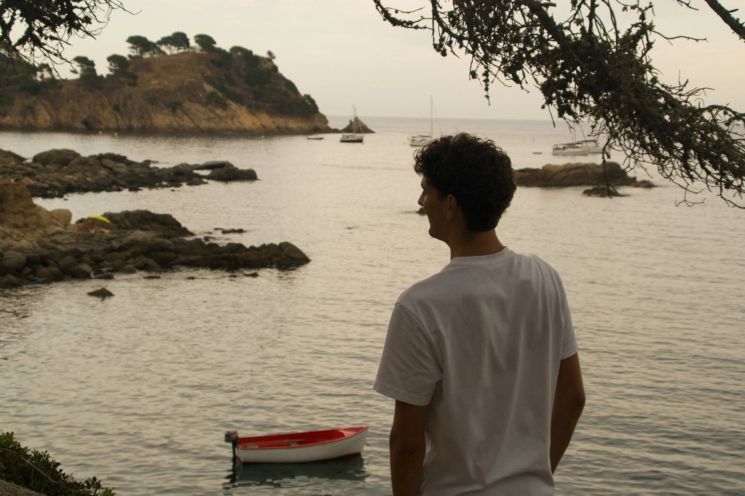 Effortless Mediterranean lifestyle portrait of a young man in a white cotton T-shirt overlooking a secluded Costa Brava bay and sailing boats.