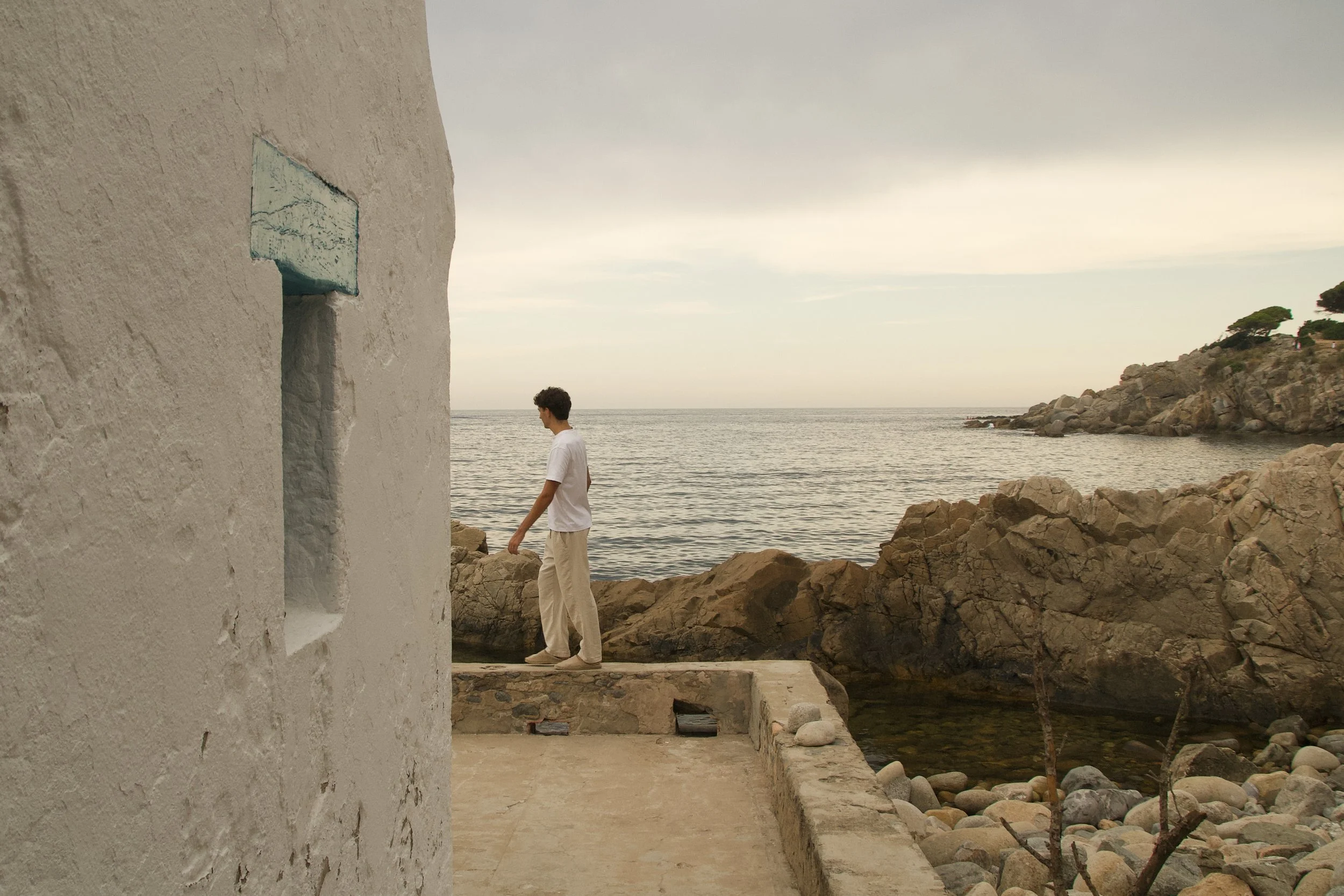 Relaxed Mediterranean fashion portrait of a young man in a white cotton T-shirt and linen trousers walking beside the sea on the Costa Brava.