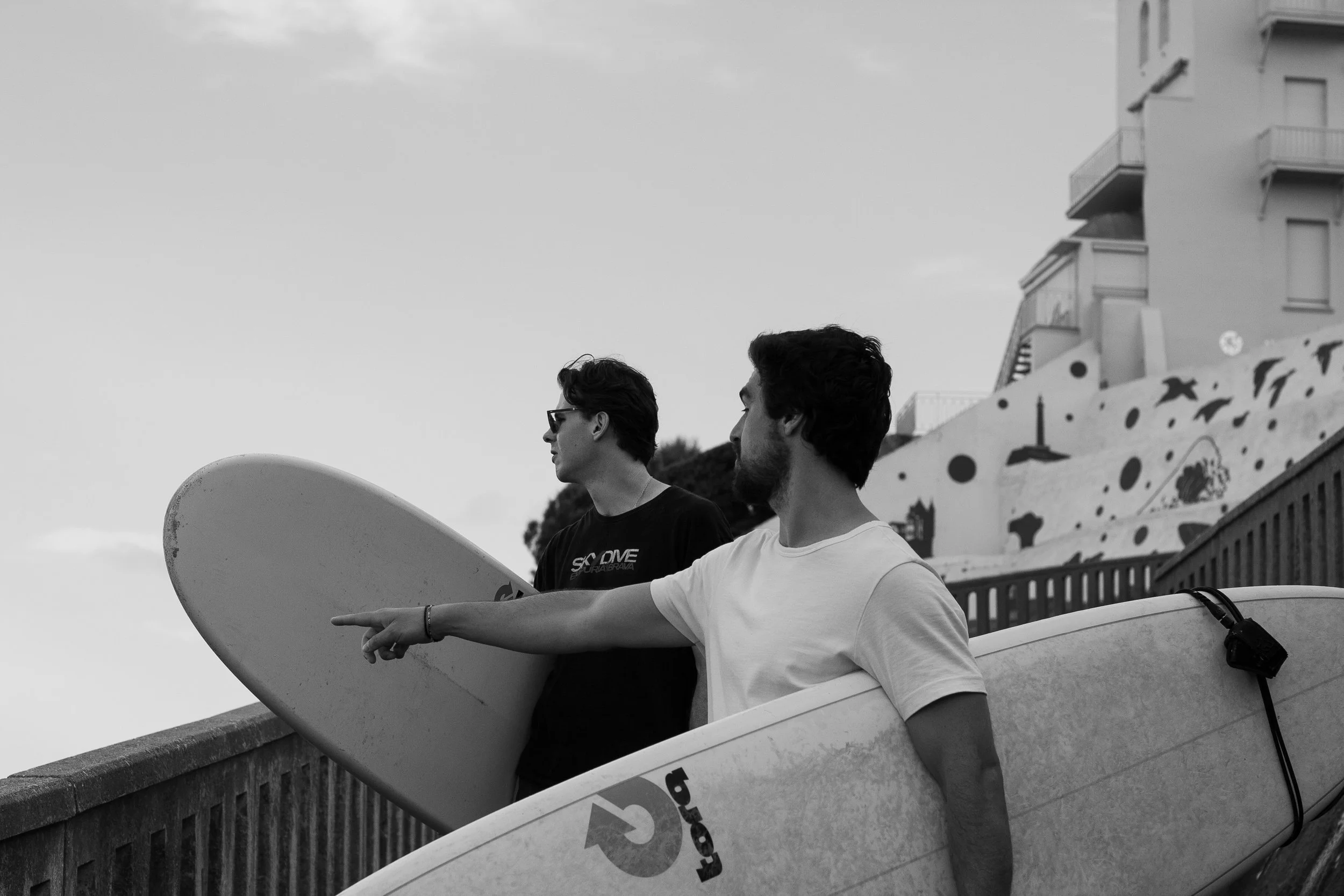 Candid street-style portrait of two surfers carrying their boards in Biarritz, photographed naturally as they pause to watch the ocean.
