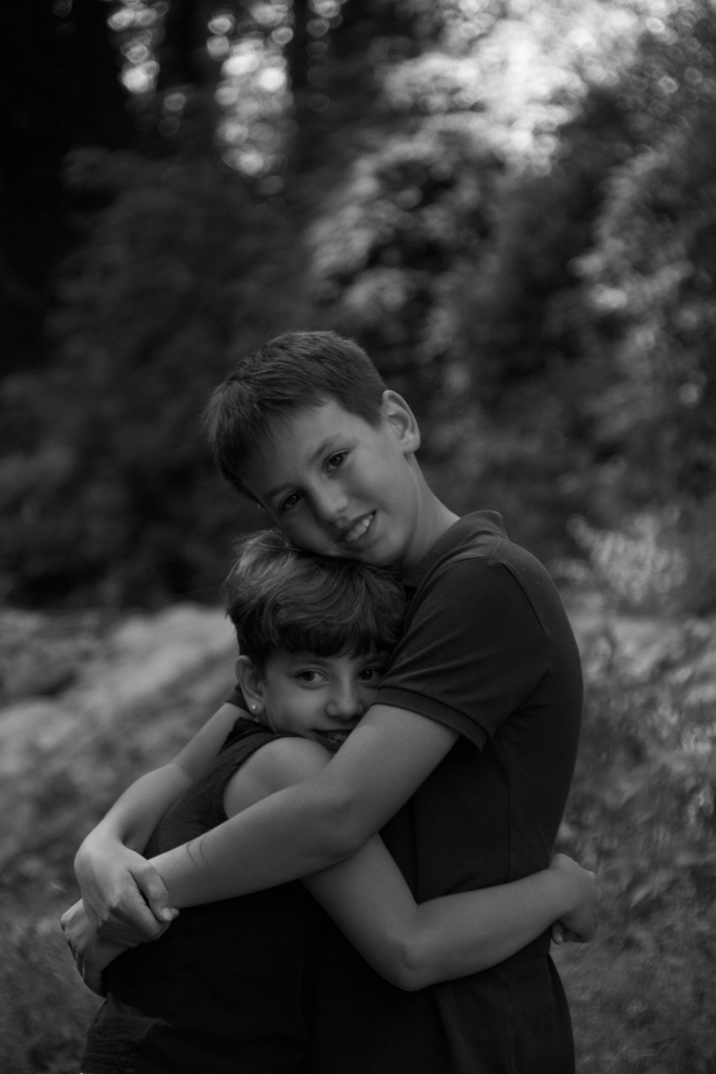 Black-and-white family portrait of two siblings embracing outdoors, capturing a quiet, affectionate and completely natural childhood moment.