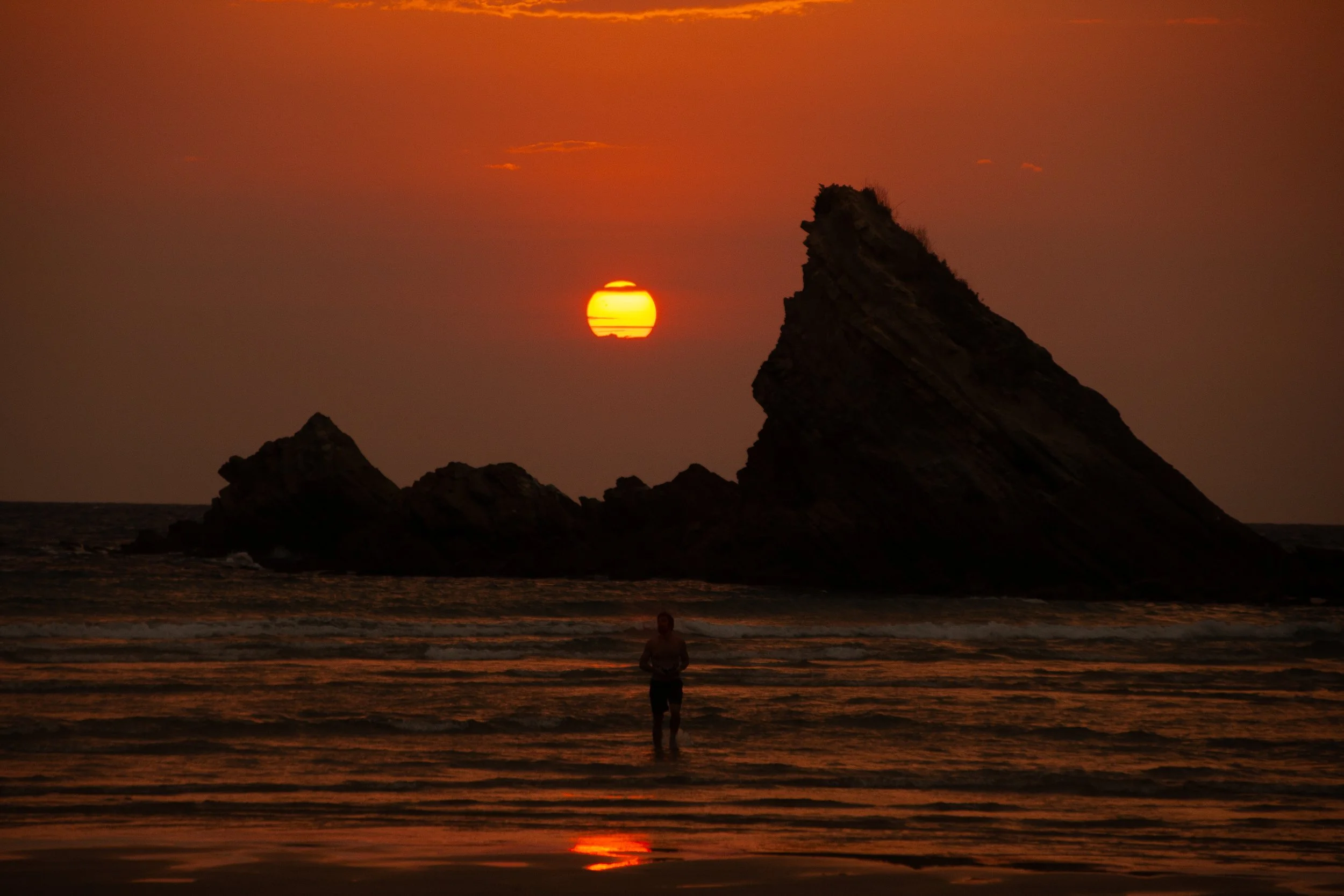 Editorial travel photograph of the Biarritz coastline at sunset, with a lone swimmer framed by dramatic rock formations and warm Atlantic light.