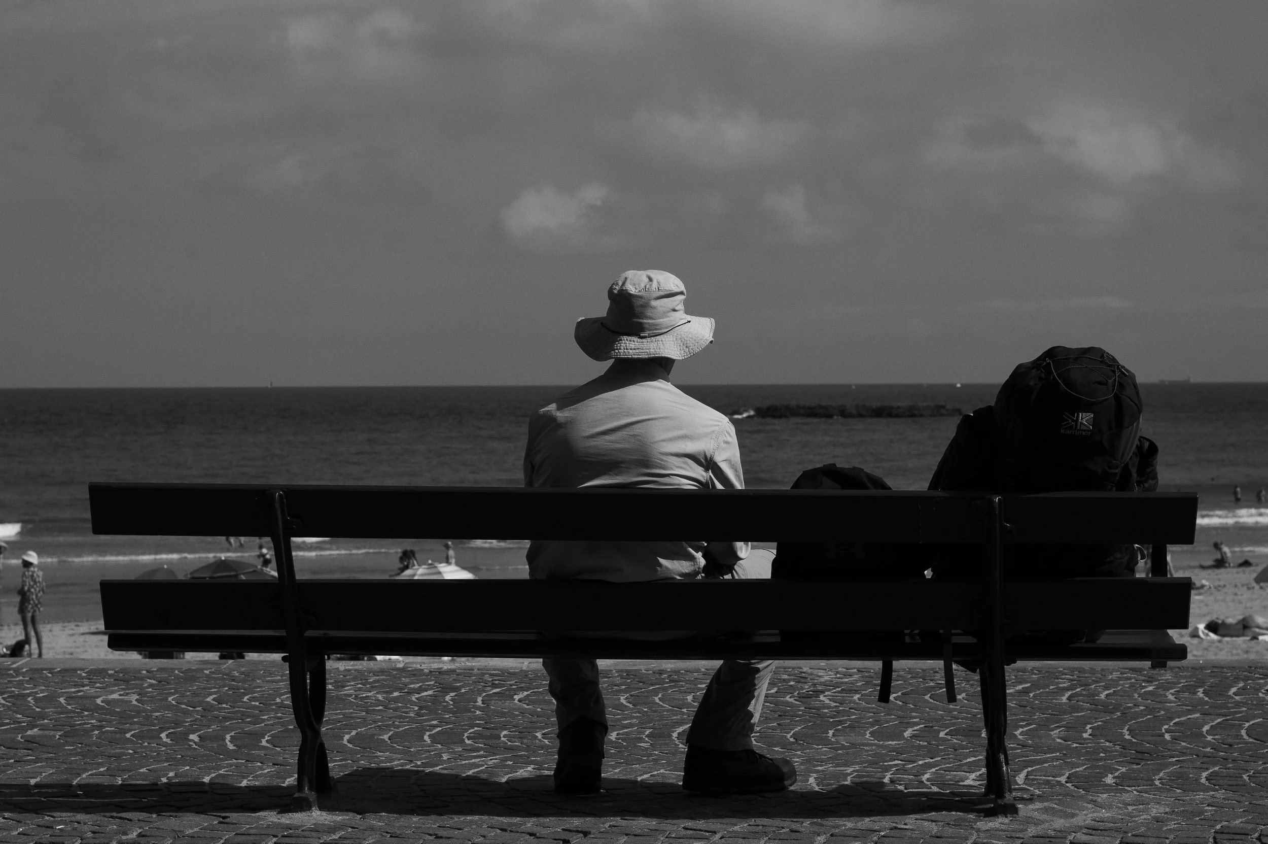 Black-and-white travel photograph of a man sitting alone on a bench overlooking the beach in Biarritz, capturing a quiet moment within everyday coastal life.