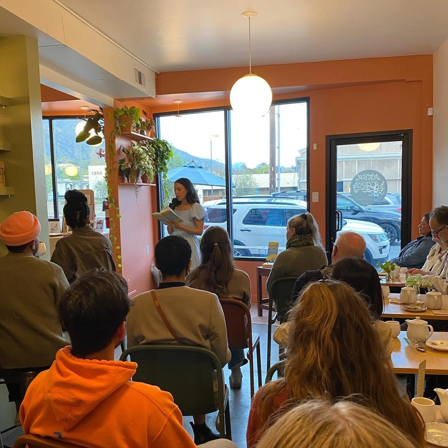 A woman standing and reading from a book or paper to a seated audience in a cozy cafe with large windows, potted plants, and warm lighting.