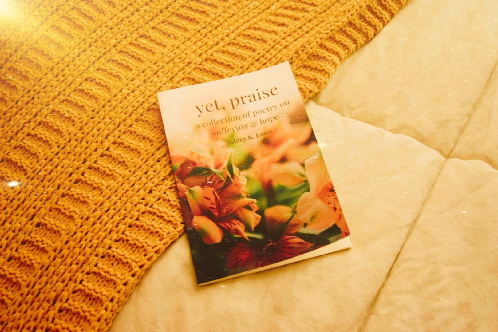 A book titled "yet, praise" resting on a yellow knit blanket next to a light-colored marble surface.