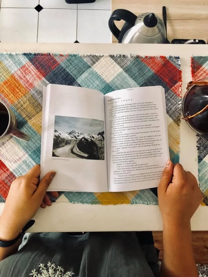 Person reading a book at a table with a colorful plaid tablecloth. The table has a cup of coffee, a pair of sunglasses, and a kettle in the background.