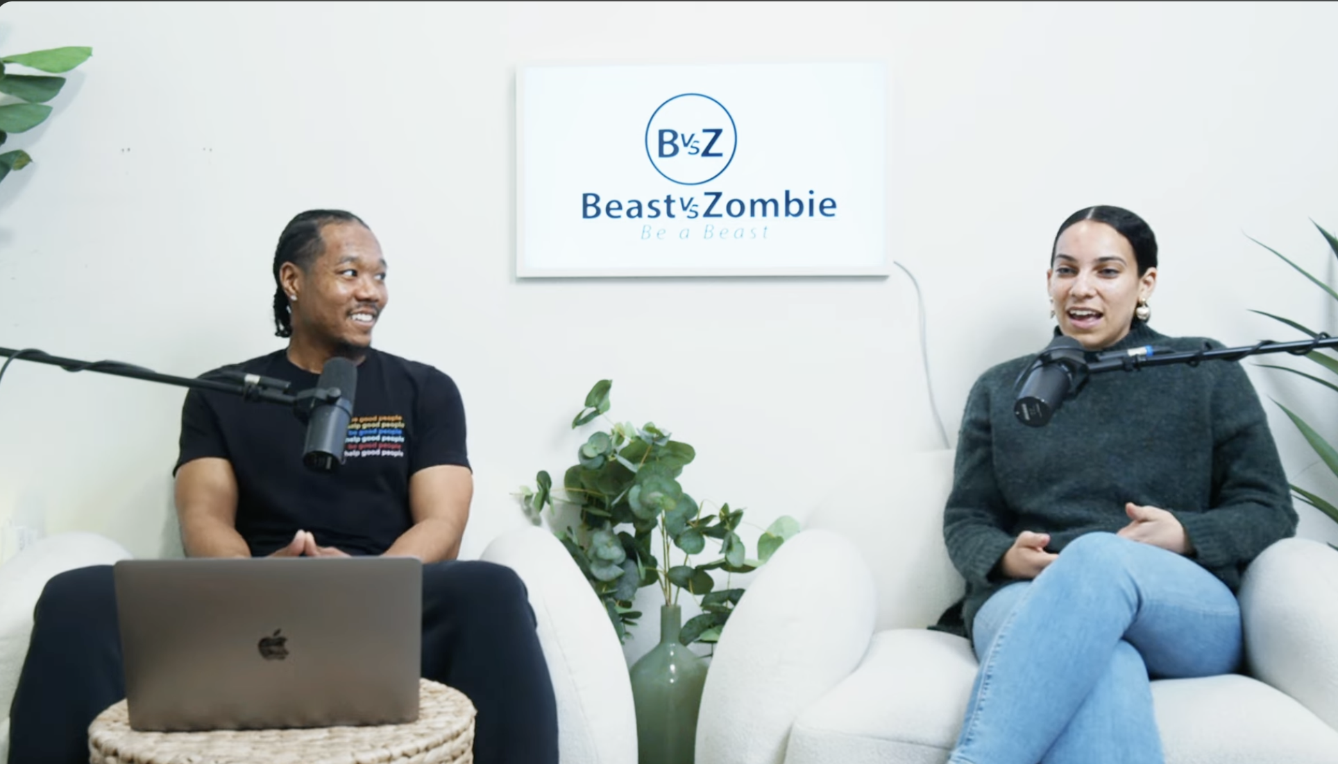Two people sitting on a white couch in a podcast studio, engaged in conversation. A sign behind them reads 'Beast vs Zombie,' with the tagline 'Be a Beast.' There are plants on either side of the sign and microphones in front of both individuals.