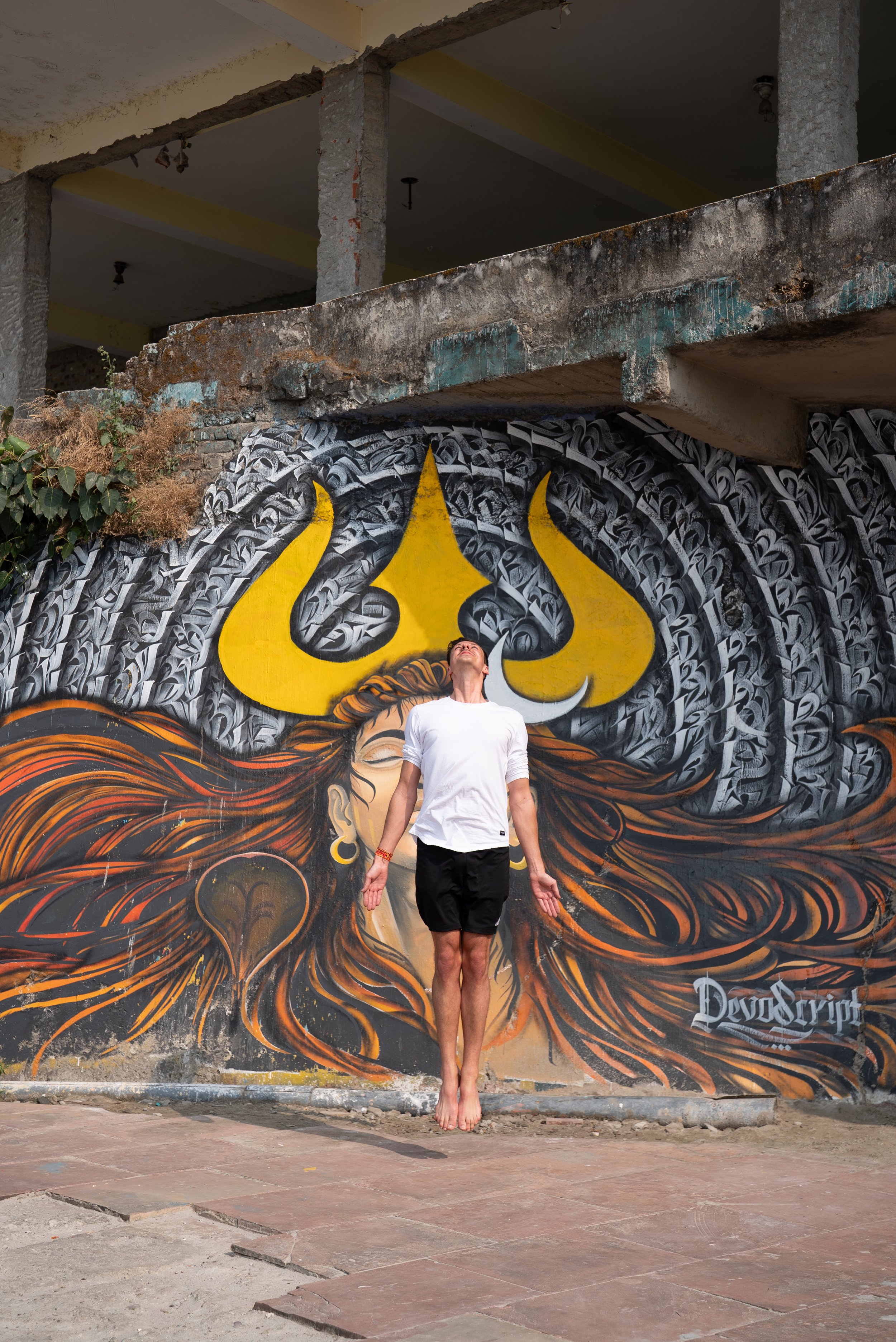Ein Mann steht vor einer Wand mit Graffiti, mit den Armen nach unten gestreckt, barfuß, mit weißen T-Shirt und schwarzen Shorts unterhalb einer Bauruine.