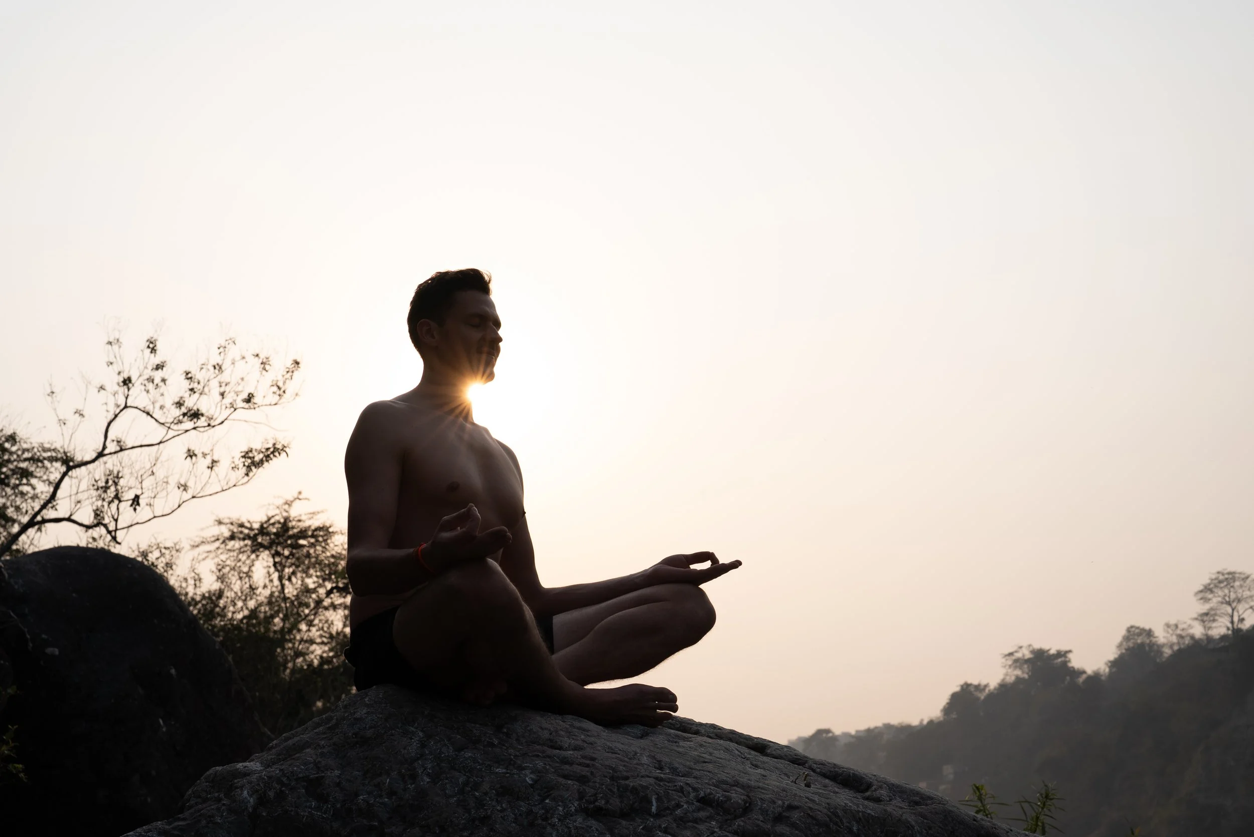 Mann in Meditation bei Sonnenuntergang mit Baum im Hintergrund.