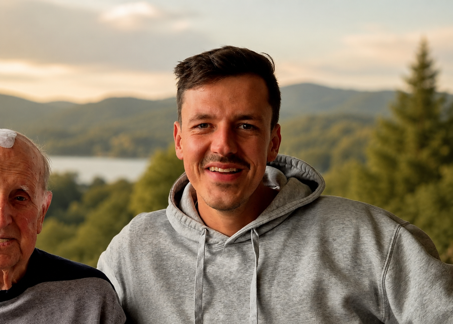 Ein junger Mann mit dunklem Haar, trägt ein graues Kapuzenpullover und lächelt in die Kamera, im Hintergrund eine natürliche Landschaft mit Wasser, Bäumen und Bergen bei Sonnenuntergang.