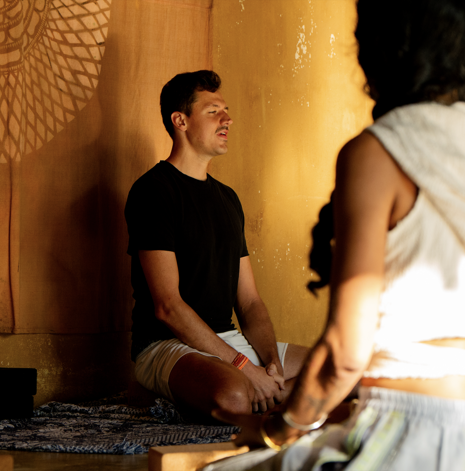 Mann sitzt auf dem Boden und unterrichtet Yoga, die Augen geschlossen, vor einer orangefarbenen Wand, während eine Person im Vordergrund zur Seite schaut.