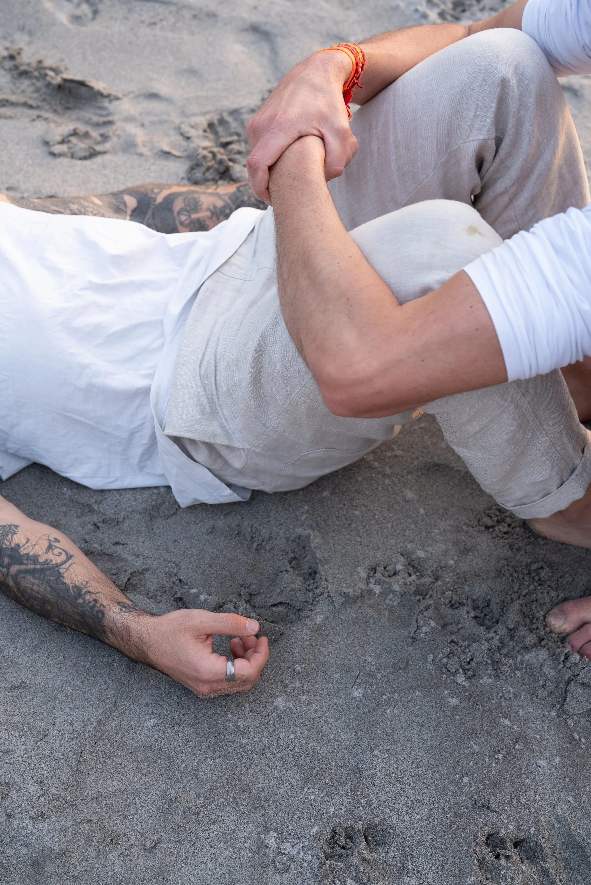 Hand mit Tattoo hält eine andere Hand auf Sandstrand, im Hintergrund eine Person mit einer Tattoos. Beide Personen tragen weiße und beige Kleidung.