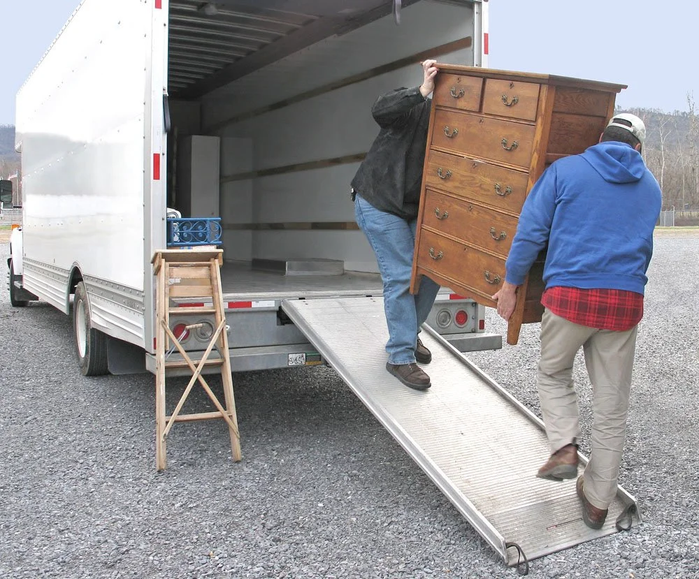 Two people unloading a wooden dresser from a truck with a ramp, outside on gravel ground.
