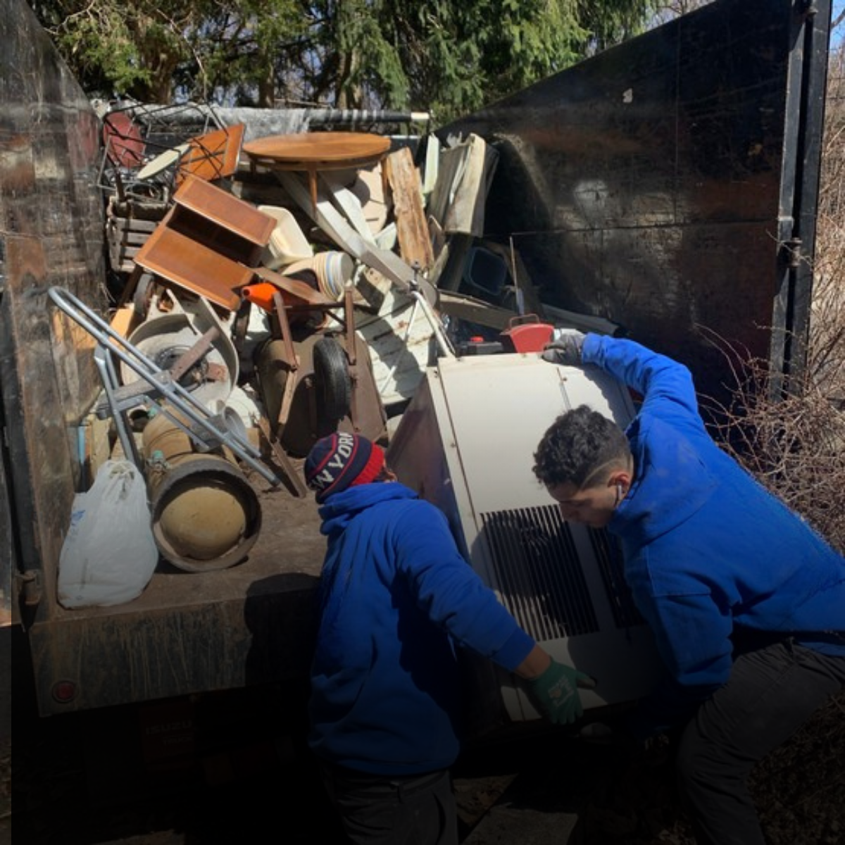 Two men in blue jackets and gloves are lifting and removing an old refrigerator from a truck filled with discarded furniture and household items, including chairs, tables, beds, and various household objects, in an outdoor area with trees in the background.