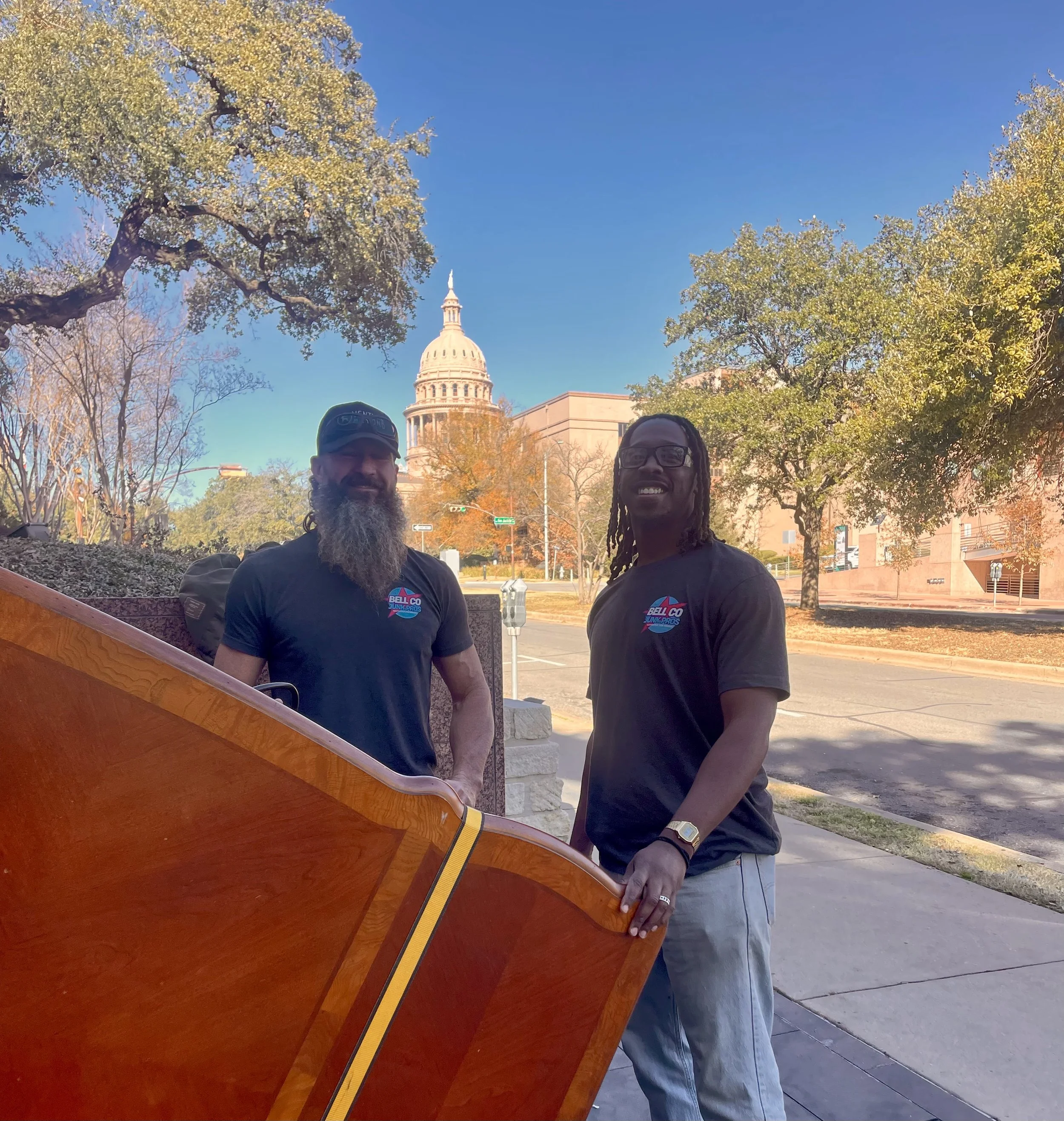 Two smiling men, one with glasses and dreadlocks and the other with a beard, stand outdoors in front of the U.S. Capitol building, with trees and a clear blue sky in the background. They are leaning on a wooden surface.